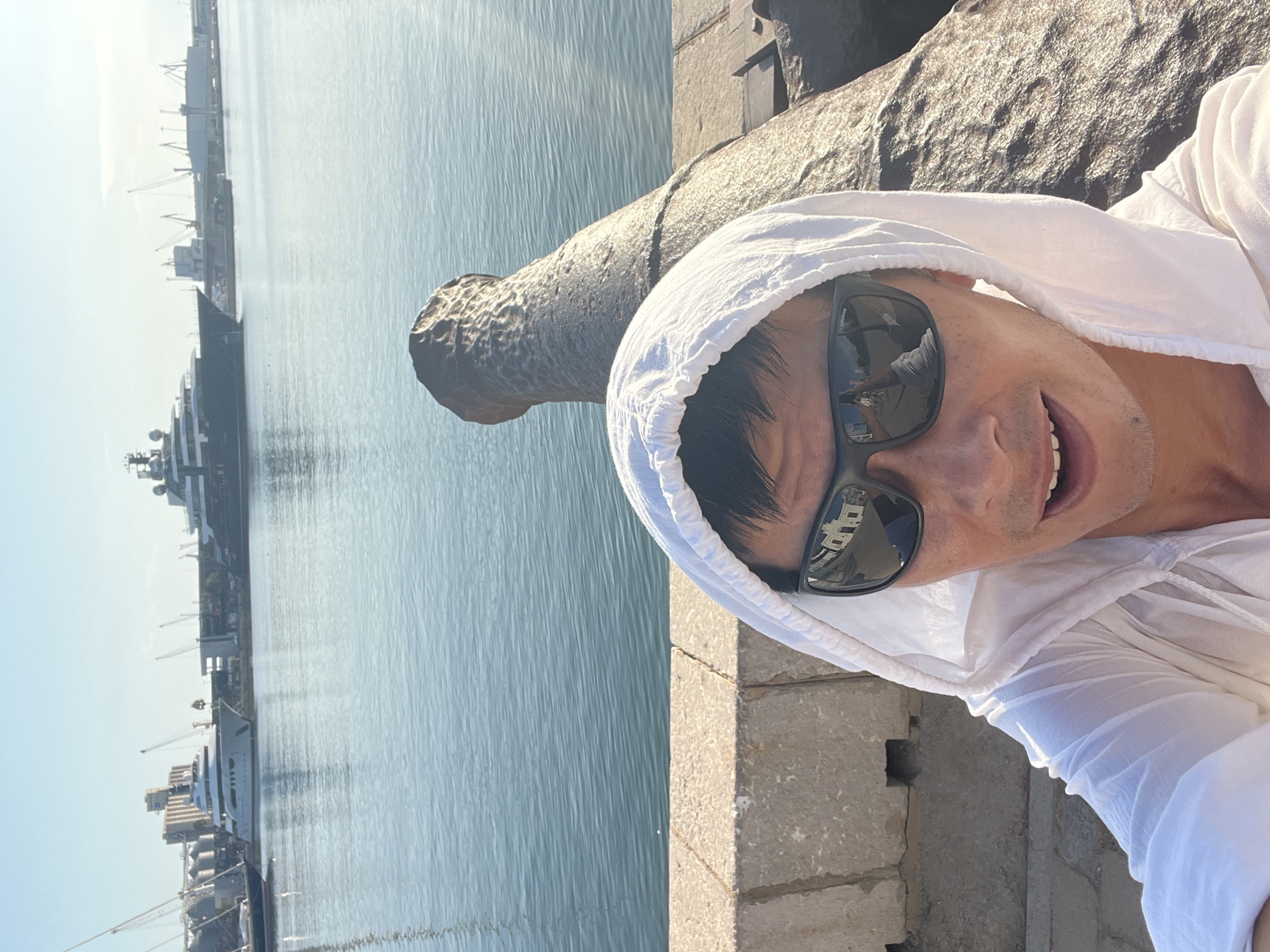 A person in a white hooded jacket and sunglasses takes a selfie in front of a large, weathered cannon. The cannon is pointed towards the water. In the background, several large yachts are docked in the harbor of Tarragona, Spain. The sky is clear and blue, and the sun is shining. The image captures a sunny day at the port, with a juxtaposition of historical elements (the cannon) and modern luxury (the yachts).