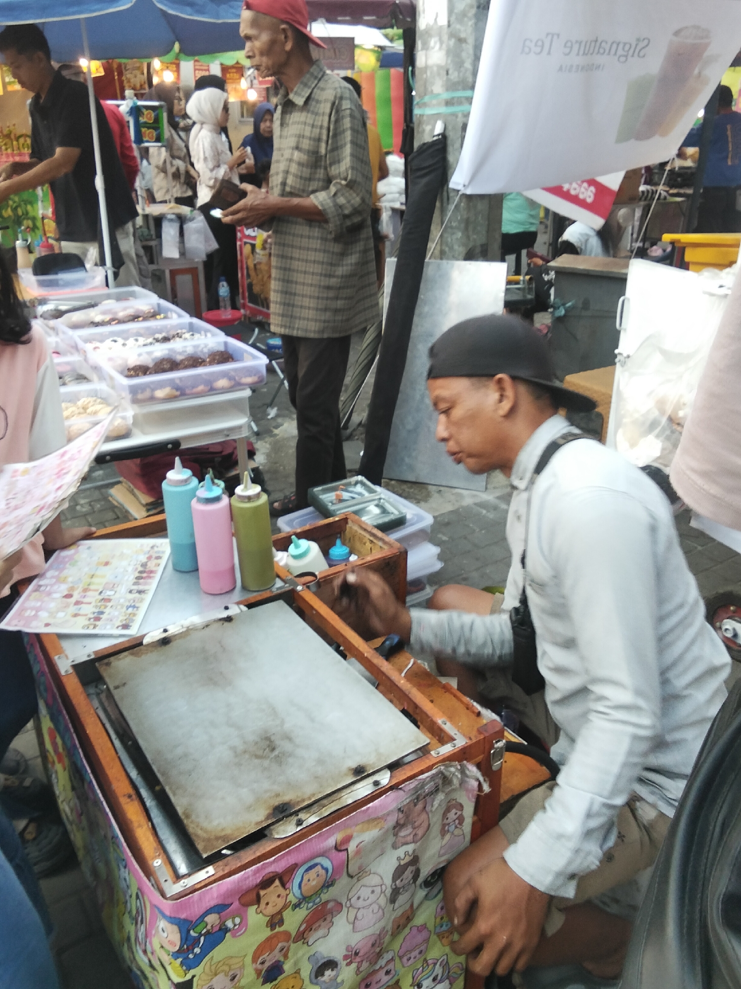 At a street market stall in Cisaat, Indonesia, a vendor, identified as a man wearing a red cap and a plaid shirt, is seen in the background behind a display of pastries. In the foreground, another man, wearing a black cap and a light-colored button-down shirt, is interacting with the pastry cart. He is seated and appears to be preparing food or serving customers. The cart itself is decorated with cartoon characters and displays several squeeze bottles of condiments. Behind the cart, a selection of donuts and pastries are neatly arranged in plastic containers. The scene suggests a typical outdoor food vending operation.