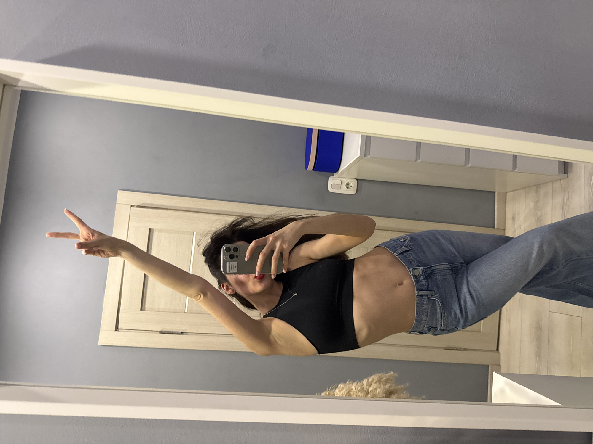 A woman is seen in a mirror selfie indoors. She is wearing a black top and blue jeans. Her left arm is raised, displaying a peace sign. She holds a smartphone in her right hand, pointed towards the mirror to capture the image. Behind her reflection is a light-colored door, a gray wall, and a wall-mounted cabinet. The scene appears to be taken in a residential setting, with the city and specific location unknown.