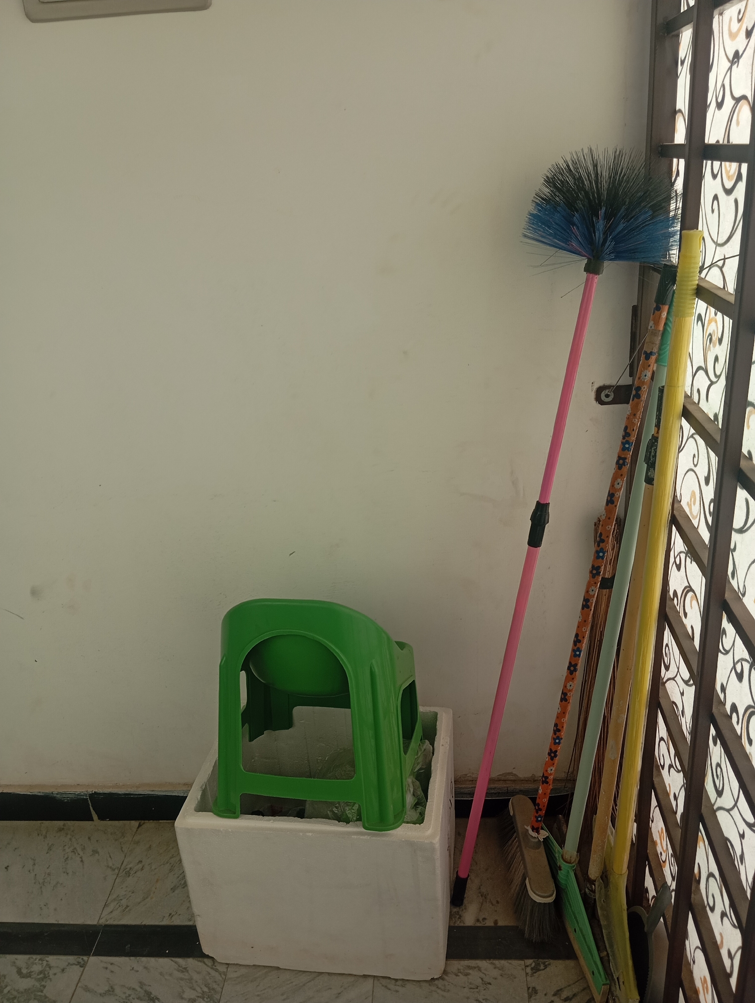 A green plastic stool is placed inside a white rectangular container next to a wall in Lassan, Pakistan. Several brooms of various colors and styles are leaning against the wall next to a barred window. The brooms have long handles and are of different sizes, with one broom having a bright blue and black brush head. The environment appears to be outdoors, possibly a patio or balcony, with a tiled floor visible. The wall is painted a light color, and the window has an ornate metal grill. No people are present in the scene.