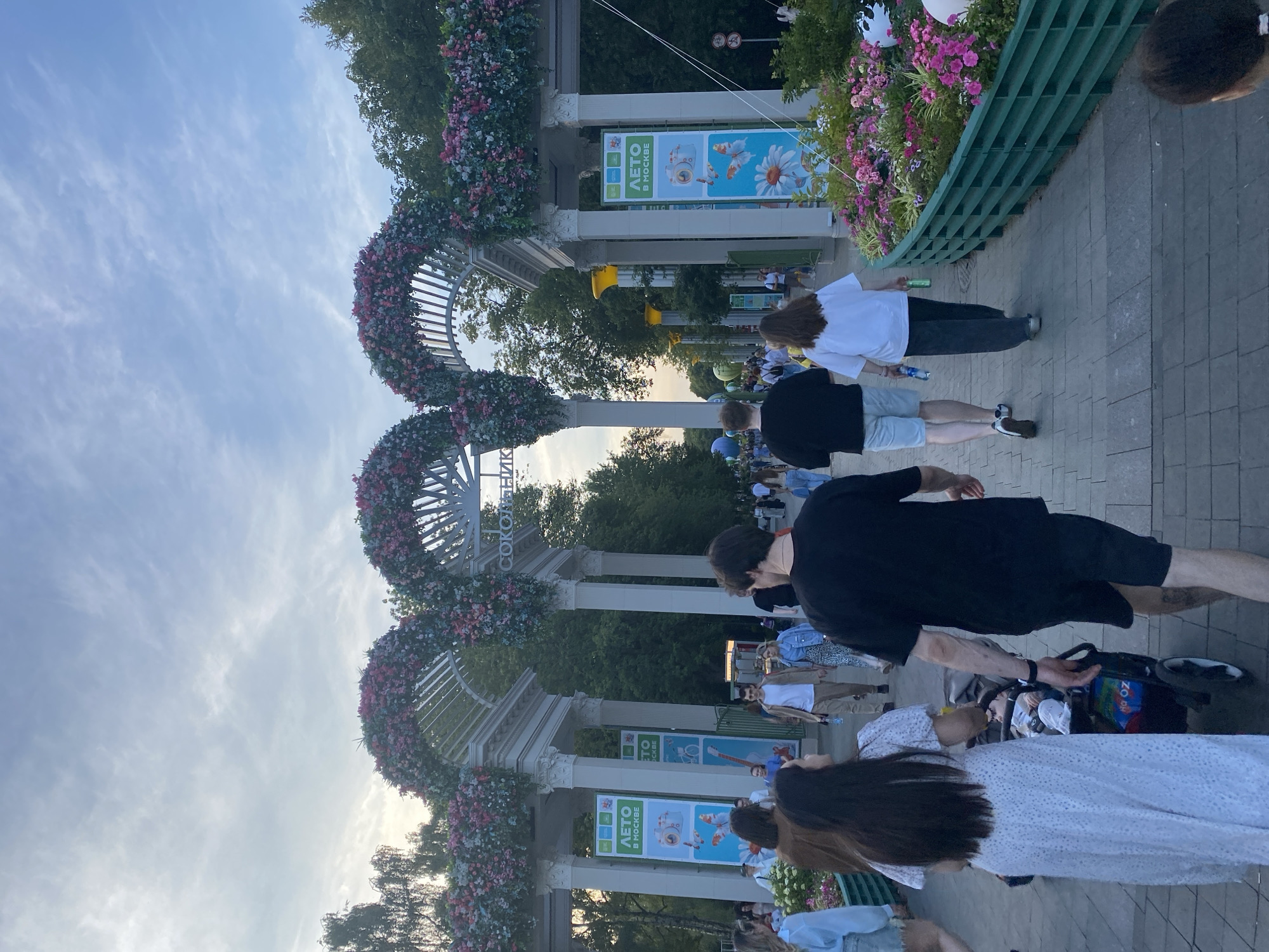 In the city of Moscow, Russia, a group of people are walking along a paved pathway lined with blooming flowers and decorative arches adorned with floral arrangements. The pathway is part of a park or public space, possibly Sokolniki Park, indicated by Cyrillic signage visible in the image. Several people are captured in motion, walking away from the camera. In the foreground, a couple is pushing a stroller, with a child visible inside. Other individuals are walking in various directions, some in casual attire, suggesting a leisurely outing. Large banners with graphics and text, including Cyrillic script, are displayed on pillars supporting the arches, contributing to the festive atmosphere. The sky above is partly cloudy, with soft daylight illuminating the scene. The overall impression is that of a vibrant public space during a pleasant time of day.