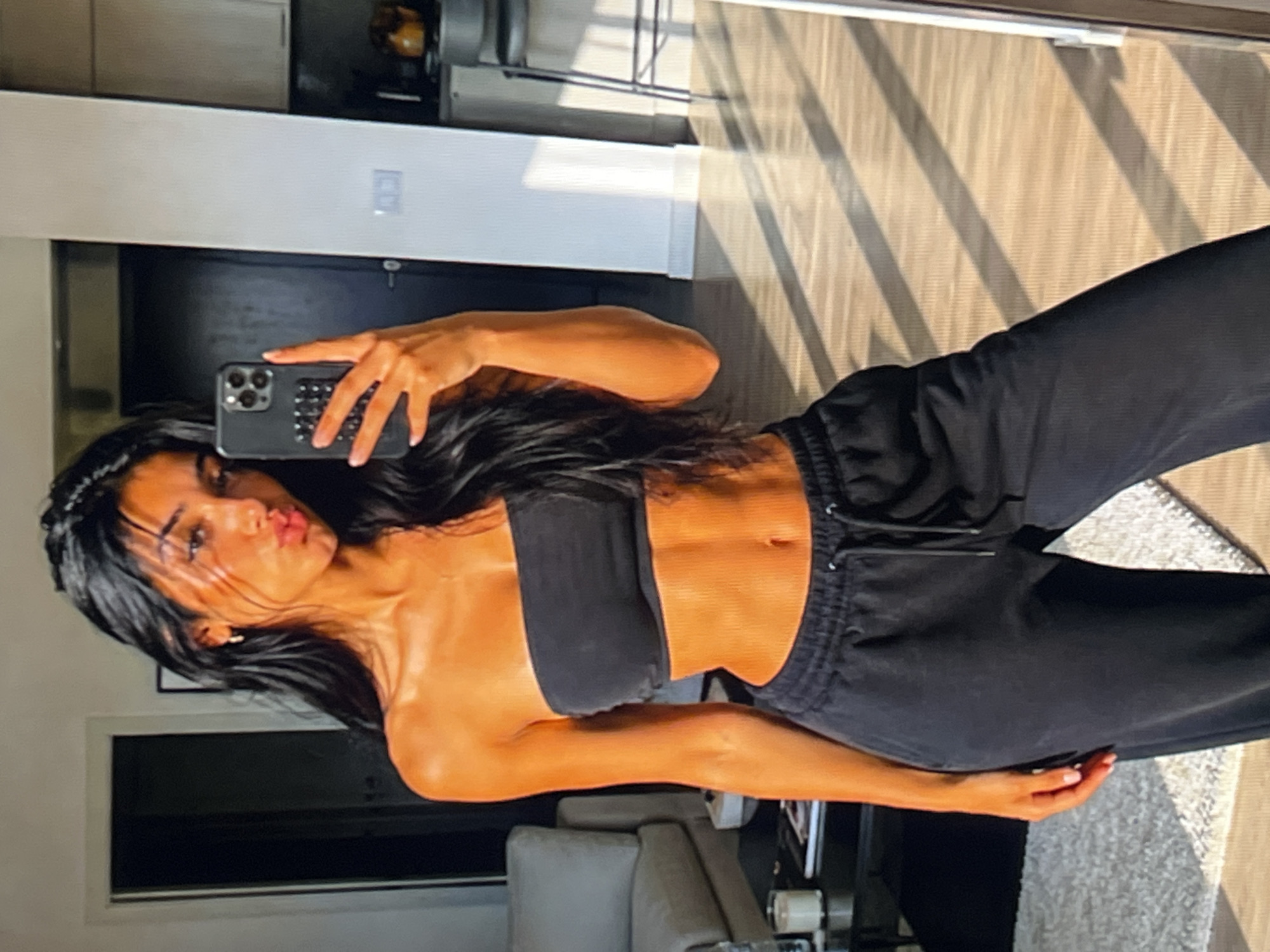 A young woman with long, dark hair takes a mirror selfie in what appears to be an apartment. She is wearing a black strapless top and black sweatpants. The woman is pouting her lips and holding a smartphone in her right hand. The background of the image shows a living room with a couch and a dining area with a bar stool. Sunlight is streaming through a window, casting diagonal shadows on the floor.