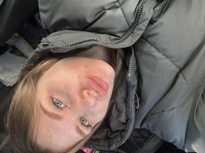 A female subject is depicted in a close-up, angled selfie, positioned inside a vehicle. She has light-colored hair, blue-green eyes with prominent eyelashes, and sculpted eyebrows. Her lips are pursed, and a small dark mark is visible below the left corner of her mouth. She is wearing a dark olive-green quilted puffer jacket with a visible zipper and a drawstring on the hood. The background reveals the dark interior of a vehicle, including a quilted surface to the upper left and a dark fabric seat to the lower right. A white plastic bag is visible in the mid-right background, bearing black text. The text on the bag indicates "АВТО-ШИНЫ" (AUTO-TIRES), a phone number "8-342-202-02-02", a website "www.avto-shini.ru", and the specific address "Пермь, ул. Промышленная, 149" (Perm, Promyshlennaya Street, 149), identifying the location as a business specializing in car tires in Perm, Russia. A yellow object is partially visible next to the white bag.