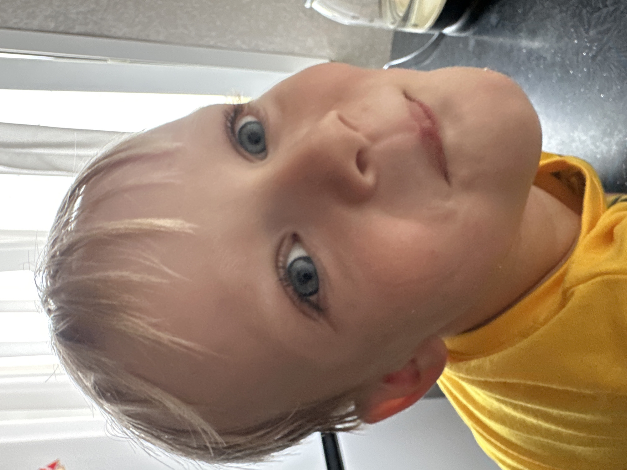 A close-up, head-and-shoulders shot captures a young boy with blond hair and blue eyes looking directly at the camera. He is wearing a bright yellow t-shirt. The background is out of focus, but appears to be an indoor setting with vertical blinds and a countertop. There is a glass container with liquid on the countertop to the right of the boy. The image was taken at an unspecified location in the city of Energetik, Russia.