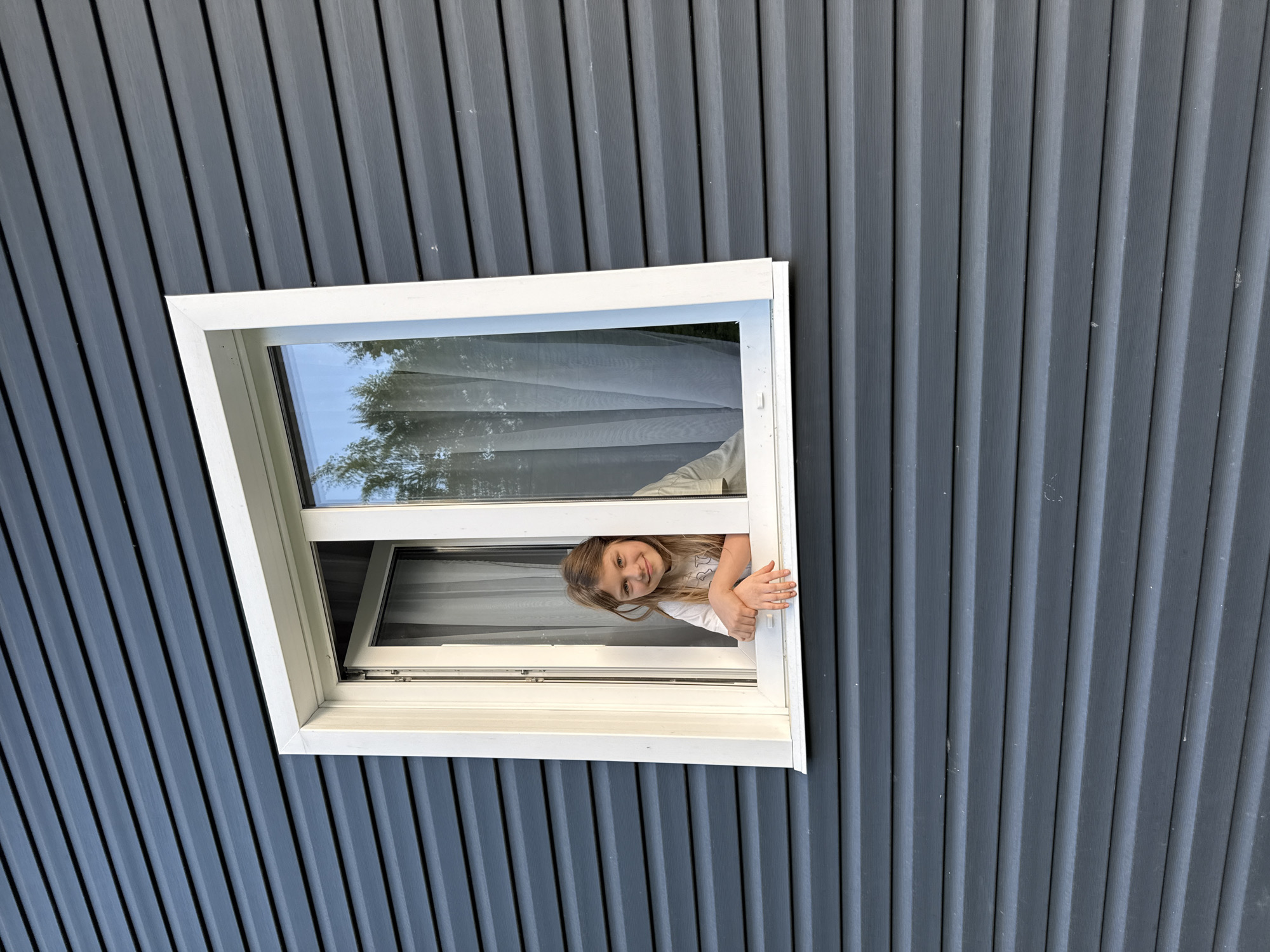 A young girl with long blonde hair is leaning out of a white-framed window in a gray corrugated metal wall. The window is open and she is smiling, resting her arms on the windowsill. Through the window, trees and a sheer white curtain can be seen. The location is likely Buzhaninovo, Russia, based on provided context.