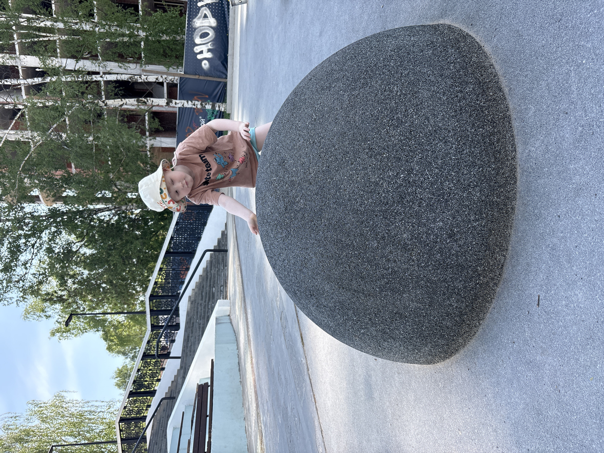 A young child in a tan shirt and patterned hat is positioned next to a large, smooth, grey rock in what appears to be a public space. The child is leaning over the rock, with one hand visible on its surface.
Behind the child, there is a building with white frames and tree branches. To the left, there are stairs with a black railing and a modern white building with wooden accents. The ground is paved with grey concrete.
Based on visual cues, this location is likely the embankment of the Izhevsk pond near the "Iron Factory" architectural complex in Izhevsk, Russia. Specifically, the stone resembles the modern art pieces and decorative stones that are placed for public interaction along the embankment, and the building in the background resembles the reconstructed facades of the Iron Factory complex.