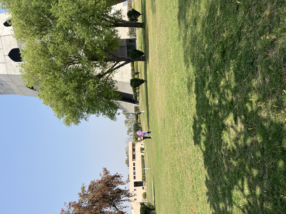 The scene is a grassy lawn in front of a large concrete structure. Two people are walking on the lawn, both wearing white shirts and pants. There are trees and shrubs in the background, and a building to the left of the scene. The sky is clear and blue. The location is Sahibzada Ajit Singh Nagar, India, specifically at the coordinates 30.704 latitude, 76.668 longitude. The image is taken from the ground looking towards the concrete structure, possibly a tower, with the two people walking towards it.