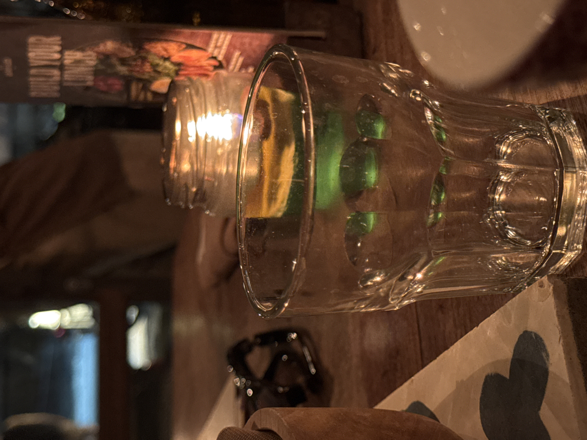 The image shows a close-up of a table setting at night, likely in a restaurant or bar in Kuta Selatan, Indonesia. In the foreground, a clear glass tumbler with a ribbed design is prominently featured. Inside the glass, a green liquid is visible, with what appear to be ice cubes or decorative elements at the bottom. To the left of the glass, a portion of a wooden bowl or container is seen, and further left, a pair of dark-rimmed glasses rests on the table. In the background, a lit candle in a glass jar casts a warm glow, illuminating a menu that displays the words "BUILD YOUR LUNCH". The overall ambiance suggests a relaxed evening setting, with soft lighting and a focus on dining and drinks.