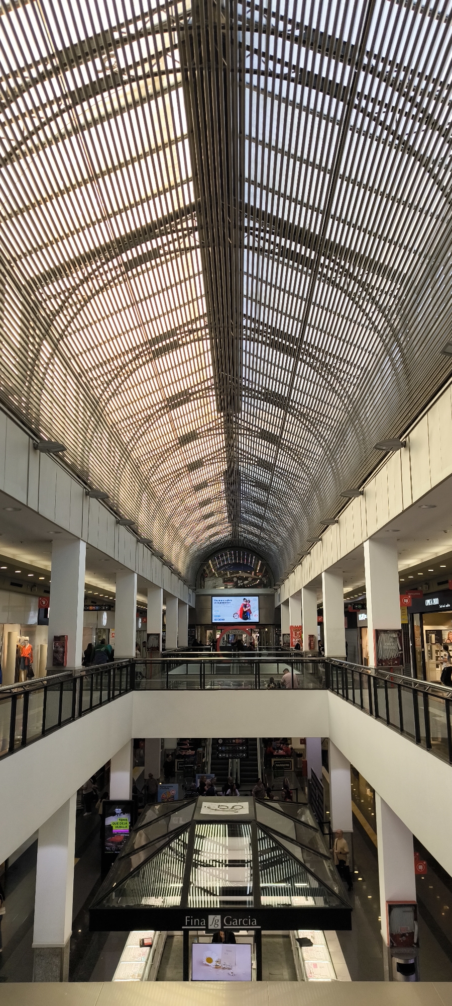This vertical shot captures the interior of a multi-level shopping mall, characterized by a striking arched, slatted skylight ceiling. The ceiling is composed of numerous parallel metal bars that create a dynamic pattern of light and shadow on the surfaces below, indicating natural daylight filtering in. The arched structure appears to have two distinct halves, possibly due to different materials or construction.
The mall features at least three visible levels. The upper levels are lined with various shopfronts and are supported by large, white rectangular columns. Black metal railings with glass panels border the open spaces, providing views down to the lower floors. Several people are visible throughout the mall, walking, standing, or using the escalators, engaging in typical shopping center activities.
At the far end of the upper level, a large digital display screen shows an advertisement. Below, prominently positioned at an entrance to a lower level, is a structure with a glass roof bearing the name "Fina fg Garcia." To its left, another vertical digital screen displays a red car and the Spanish text "TENIS QUE DEJA HUELLA." The overall environment is modern and well-lit, consistent with a contemporary shopping complex.