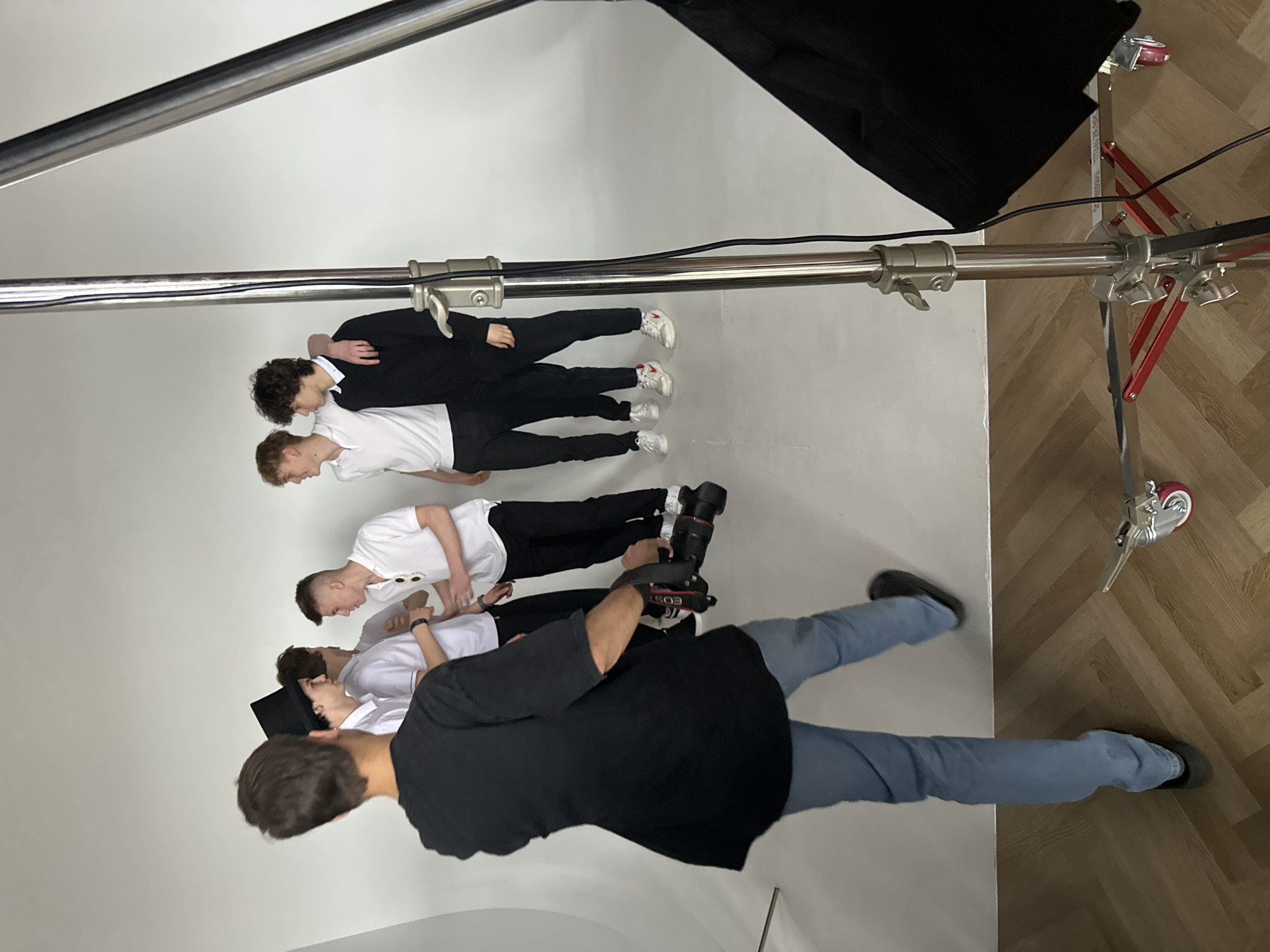 The image shows a behind-the-scenes view of a photo shoot in an indoor studio, located in the city of Unknown, Unknown. Four young men, all wearing variations of white shirts and black pants, are lying horizontally on a white backdrop. The man closest to the camera is taking a picture of the group. He is wearing a black t-shirt and blue jeans, and is holding a DSLR camera. Overhead, a silver boom arm supports a black rectangular light modifier. The base of the boom is a red and silver stand with wheels. The floor in the foreground appears to be wood.