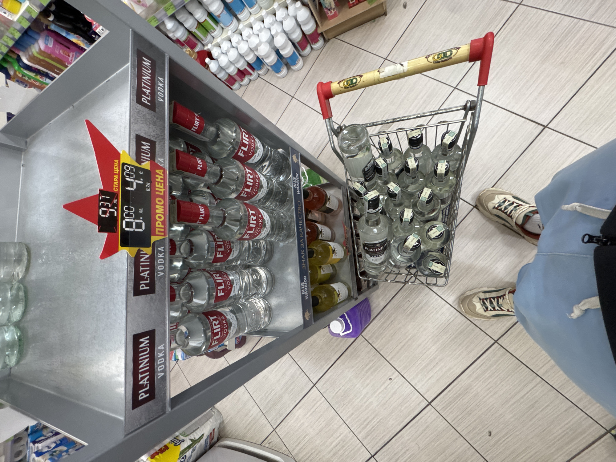 The image shows a shopping cart in a store aisle, filled with bottles of "Platinum Vodka." The bottles are positioned next to a shelf with "Flirt Vodka," with a price display indicating a discount from 9.37 to 8.09, presumably Bulgarian lev (лв.). A person wearing light blue sweatpants and beige sneakers stands next to the cart. It's a store environment based on the products and the shopping cart and there's a promotional price for the vodka, this is most likely in the city of Pleven, Bulgaria.