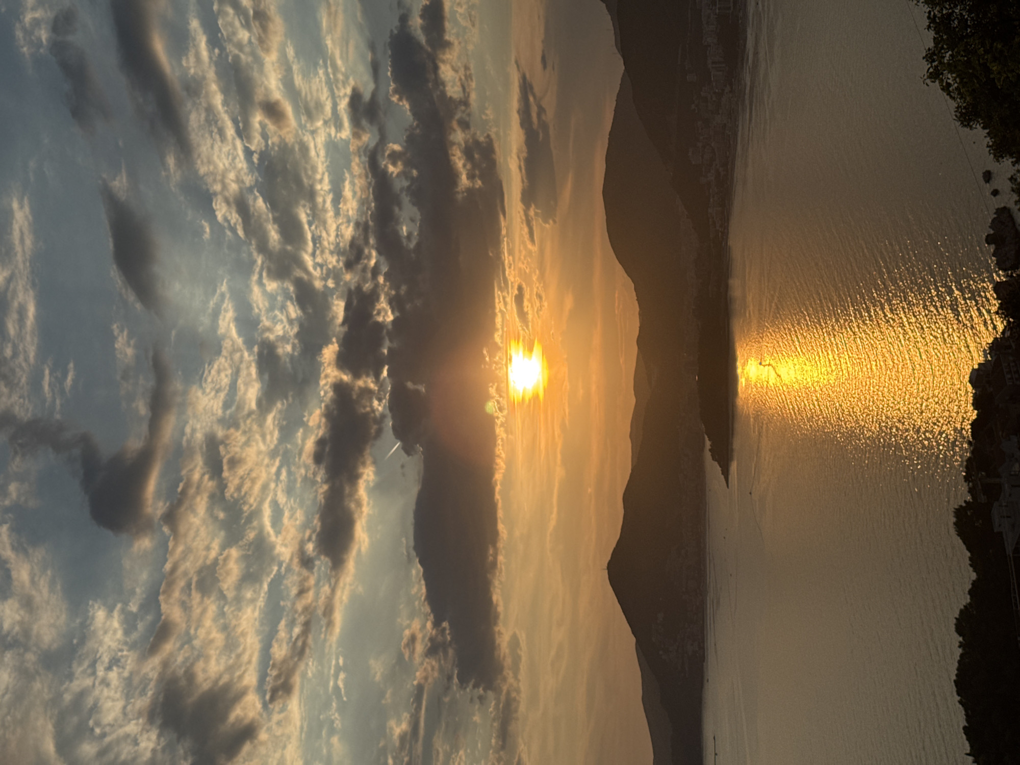 The image depicts a serene coastal landscape at sunset. The sun, a bright orb of golden light, is partially obscured by dramatic, textured clouds that stretch across the sky, casting an array of warm oranges, yellows, and blues. A vibrant reflection of the sun shimmers across the calm waters of a bay, creating a dazzling path of light. In the background, silhouetted mountains rise, their peaks softened by the twilight. Along the foothills of the mountains, a town or city is visible, its lights beginning to twinkle as dusk settles. Lush greenery, a dense line of trees, lines the foreground, framing the view of the bay and the distant settlement. The overall impression is one of natural beauty and tranquility as day transitions into night.