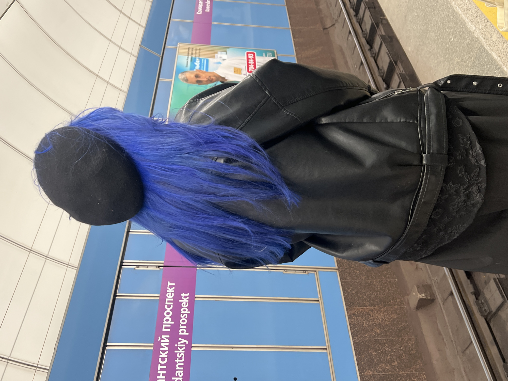 From a rear perspective, a person with vibrant, electric blue hair, partially covered by a black beret or flat cap, stands on a modern subway platform. They are wearing a black, leather-look jacket, which features a subtle patterned or embroidered detail on the lower back. The jacket has a zippered back and belt loops.
The setting is a clean, contemporary subway station, identified by signs displaying "Комендантский проспект" (Komendantskiy prospekt) in Cyrillic and "Komendantskiy prospekt" in Latin script, confirming its location in Saint Petersburg, Russia. The station walls are composed of reflective white panels intersected by blue horizontal lines.
To the right, the edge of the platform and subway tracks are visible, indicating an active railway. In the background, an advertisement billboard on the station wall shows a man in a white shirt. The billboard prominently features a phone number "704-84-93" and some partially obscured Russian text.
The person appears to be standing still, possibly waiting for a train. The indoor environment means no outdoor weather conditions are visible, and the time of day is not discernible. No specific emotions are conveyed from this rear view.