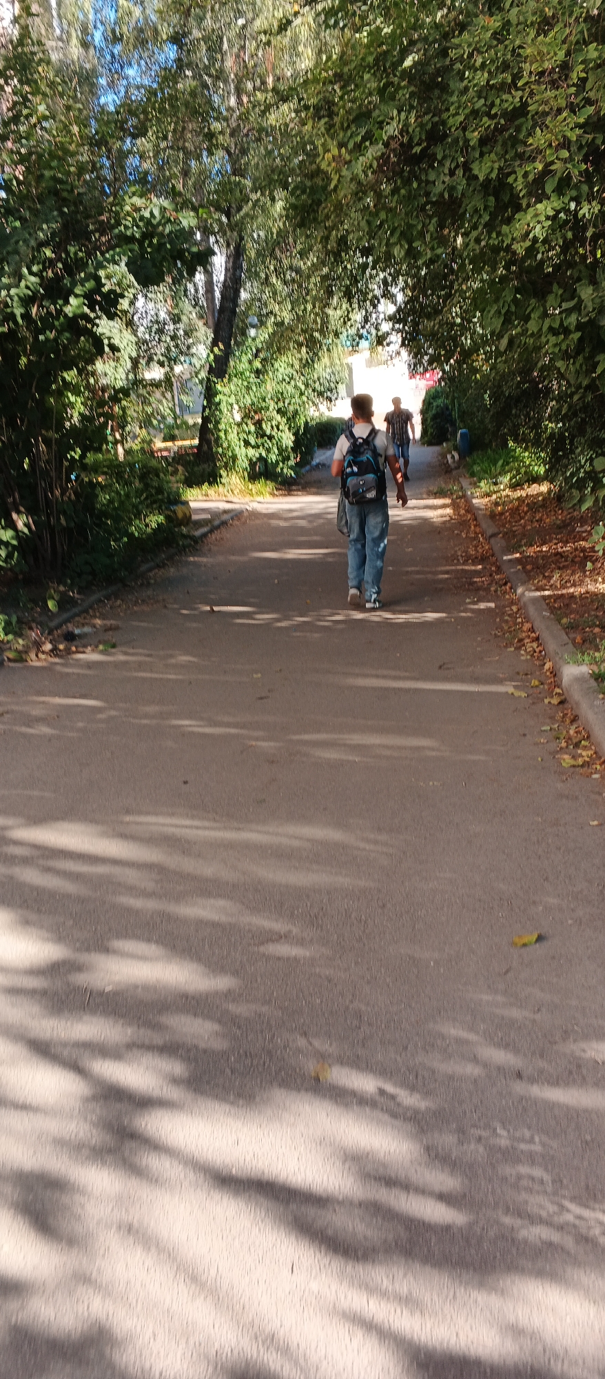 In Smila, Ukraine, a young person walks away from the camera on a paved path lined with green trees and foliage. The person is wearing a white t-shirt, blue jeans, and a dark backpack. Another person is visible further down the path, also walking away from the camera. The path is dappled with shadows from the trees. The background shows more greenery and a glimpse of buildings.