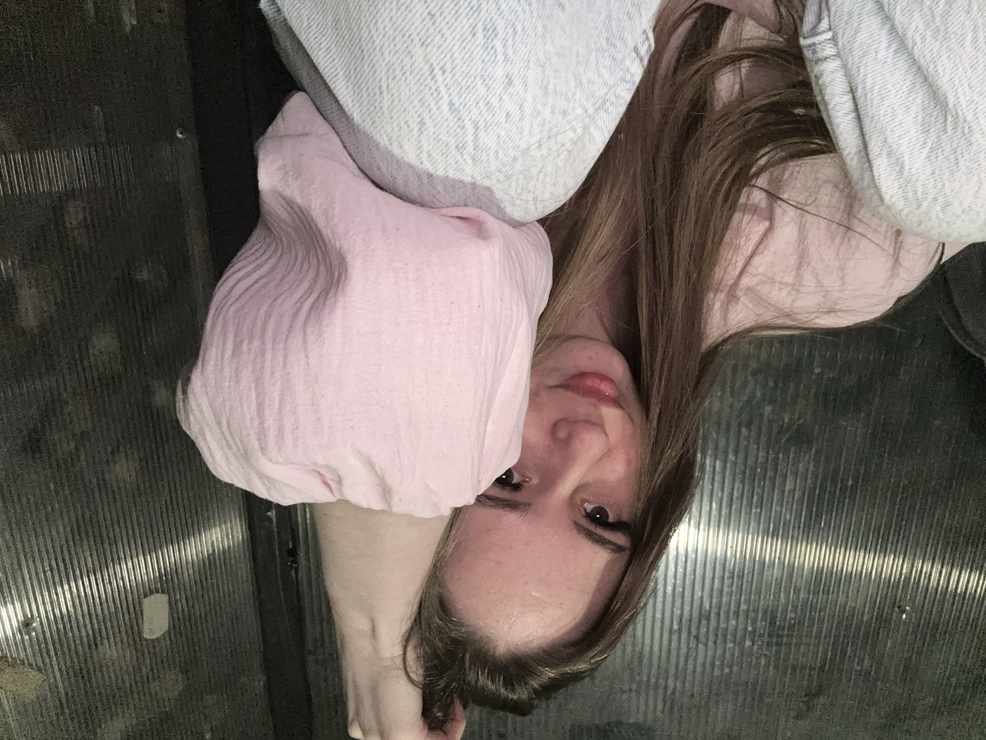 The image shows a young woman lying down or leaning against a dark, corrugated metallic surface, looking directly at the camera. Her body is oriented such that she appears upside down, with her head towards the bottom center of the frame. She has long, straight brown hair, which partially covers her face.
The woman is wearing a light pink top or garment, which has a visible texture, and light blue denim fabric, likely part of jeans or a jacket, is visible near her head and shoulder. Her face is visible, showing a neutral expression with a slight hint of a smile, and she has eye makeup, including eyeliner or mascara. Her lips are slightly parted.
The setting appears to be an indoor space with a dark, ribbed, metal surface dominating the background. This surface could be a floor, wall, or ceiling, possibly in an elevator, a utilitarian area, or an industrial-style location. Reflections of light are visible on the metallic ridges, with one notable bright, horizontal streak across the corrugated pattern. The overall lighting is dim. There is no visible text in the image.