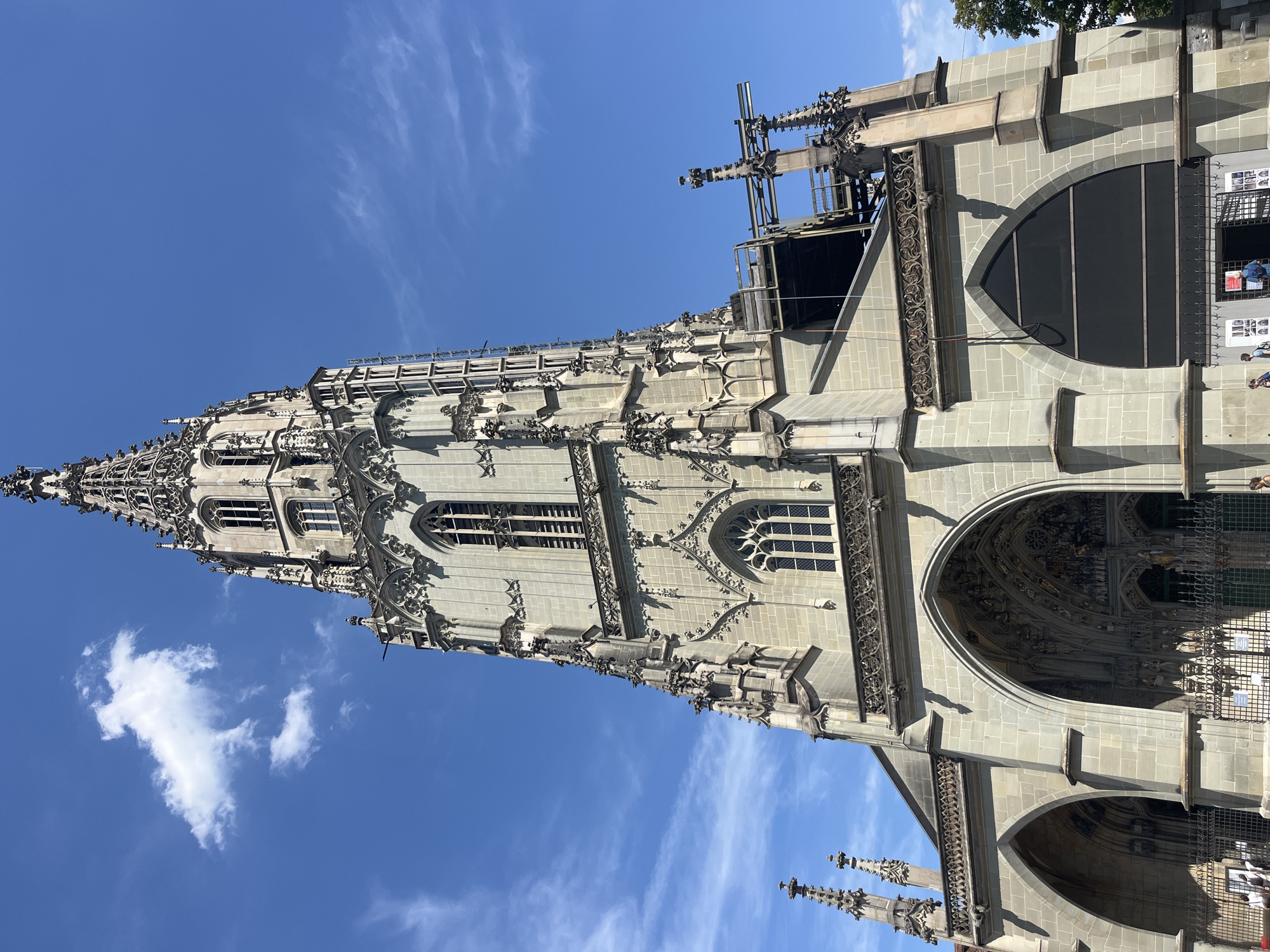 The image shows the Bern Minster (Berner Münster), a Gothic cathedral located in the city of Bern, Switzerland. The cathedral's tall, ornate spire dominates the frame, reaching towards a bright blue sky with scattered white clouds. The facade of the cathedral, constructed from light-colored stone, features intricate carvings, pointed arches, and numerous windows. Scaffolding is visible around a section of the upper part of the tower, suggesting ongoing construction or maintenance work. In the foreground, the cathedral's main entrance is visible, with large arched doorways and decorative sculptures. A black metal fence encloses the area in front of the entrance, and a few people can be seen walking near the fence. Trees are visible on the right side of the image.