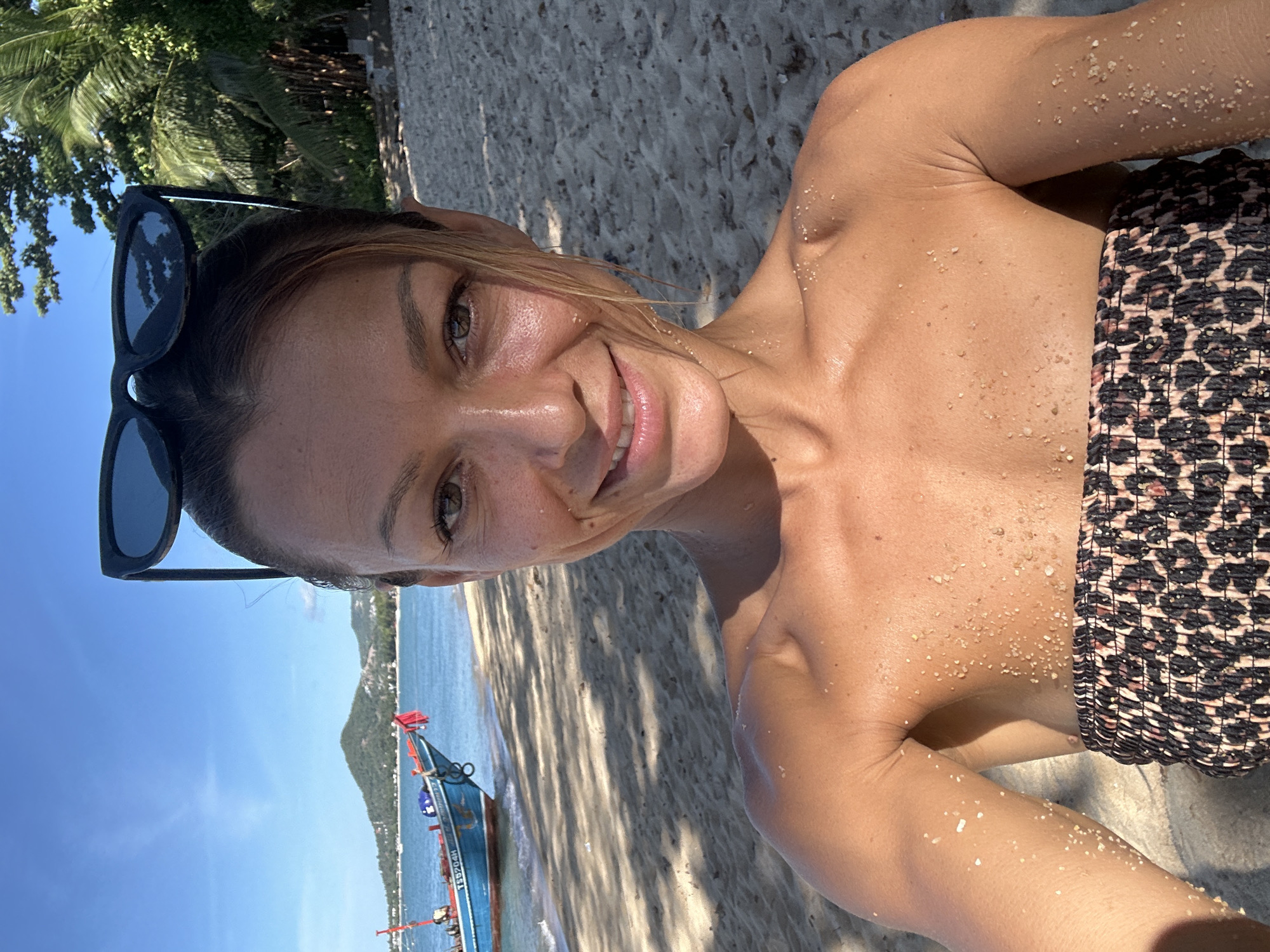 A young woman with light skin and brown hair pulled back, wearing a strapless leopard print top, is taking a selfie on a sandy beach. She has sunglasses perched on top of her head and her skin is glistening with moisture and specks of sand. The background features a blue ocean, a distant green hill, a blue and red fishing boat, and lush green palm trees under a clear blue sky. The overall scene suggests a sunny day at the beach in Baan Tai, Thailand.