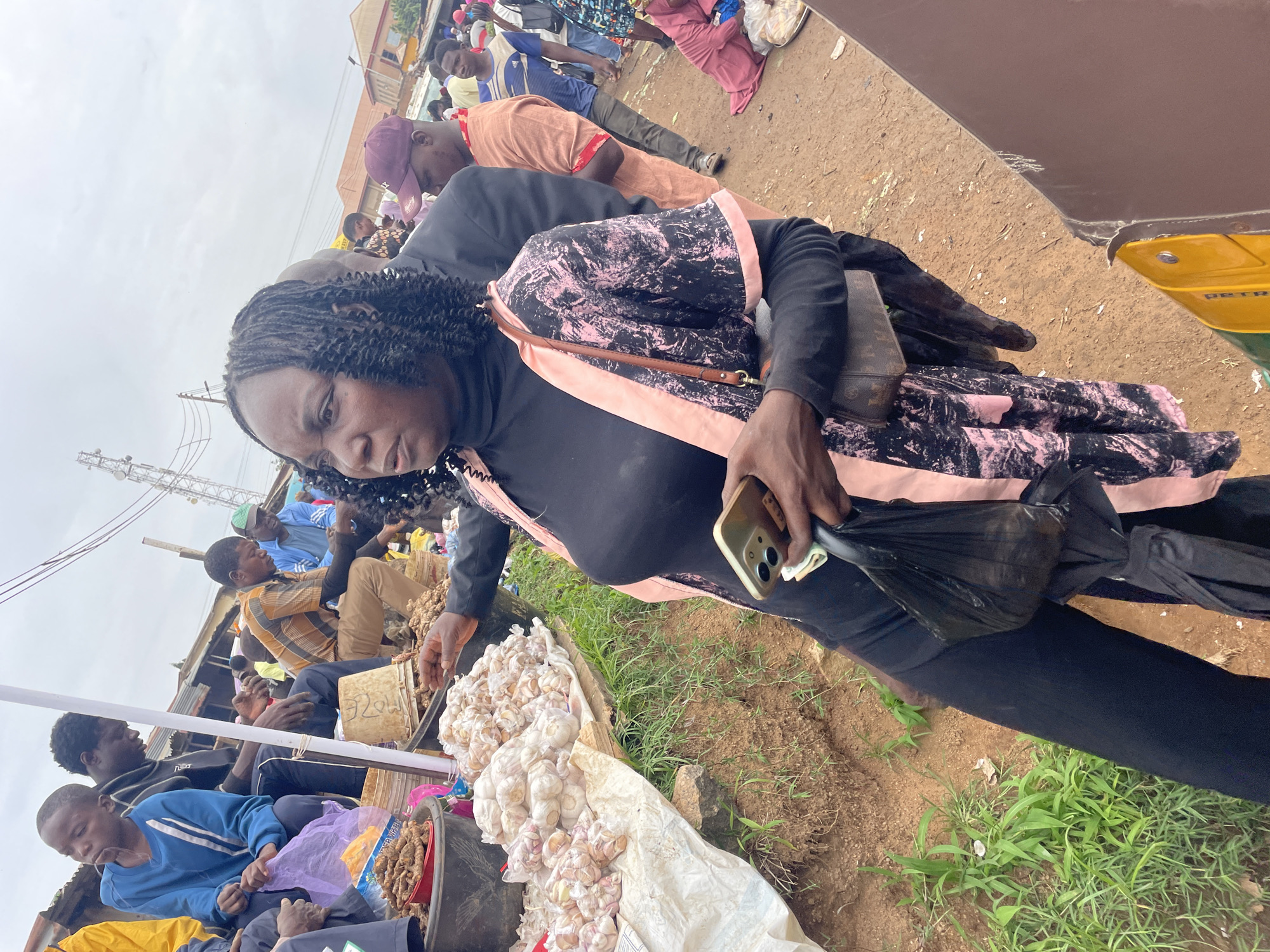 The image depicts a bustling outdoor market scene in Gura Top, Nigeria, on an overcast day. A woman in the foreground, wearing a black turtleneck, a black and pink patterned overshirt, and black pants, is holding a smartphone in her right hand and a black plastic bag with an umbrella inside in her left. She appears to be interacting with a stall or another person off-camera to her left.
To her left, a table displays a large quantity of garlic bulbs packaged in clear plastic bags. Behind the garlic, a person wearing a yellow jacket is visible. Further back, men and women are seen browsing or working at various stalls. One man in a plaid shirt is sitting near a bucket, and another man in a blue shirt is standing behind him. In the background, there are various structures, including what appears to be market stalls and buildings, with a cell tower visible on the right side of the frame. The ground is dirt and grass.