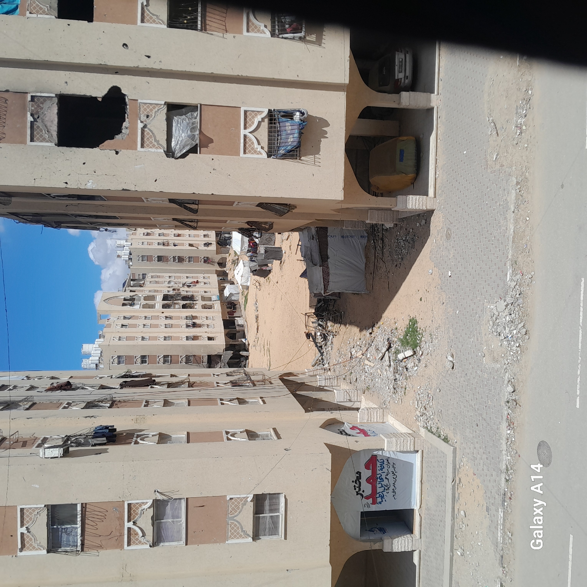 The image shows a residential area in Unknown, Unknown, likely after a conflict or disaster. Buildings display significant damage, including gaping holes in walls and windows. Tents are set up in the space between the buildings, suggesting displacement of residents. Debris litters the ground, and a sign in Arabic indicates a medical facility. The scene appears to depict a crisis situation in an urban environment.
