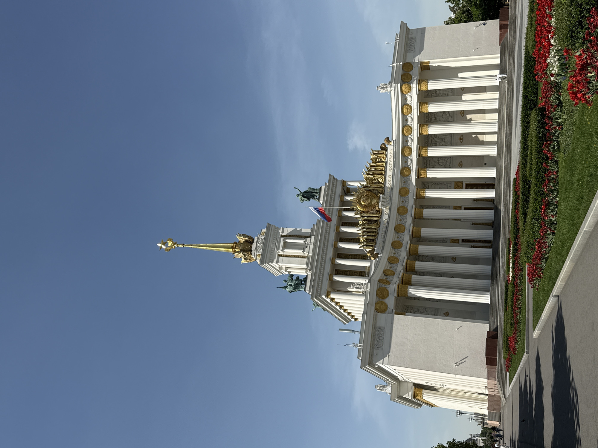 The image shows a building in Moscow, Russia, likely the "Friendship of Nations" pavilion at the Exhibition of Achievements of National Economy (VDNKh). The building features white walls, gold ornamentation, and numerous columns. A spire topped with a star rises from the center. A Russian flag is visible near the top. The sky is clear and blue. The building sits beside a walkway, with grass and red flowers visible along its edge. The angle of the photo is upward, taken from a low vantage point.