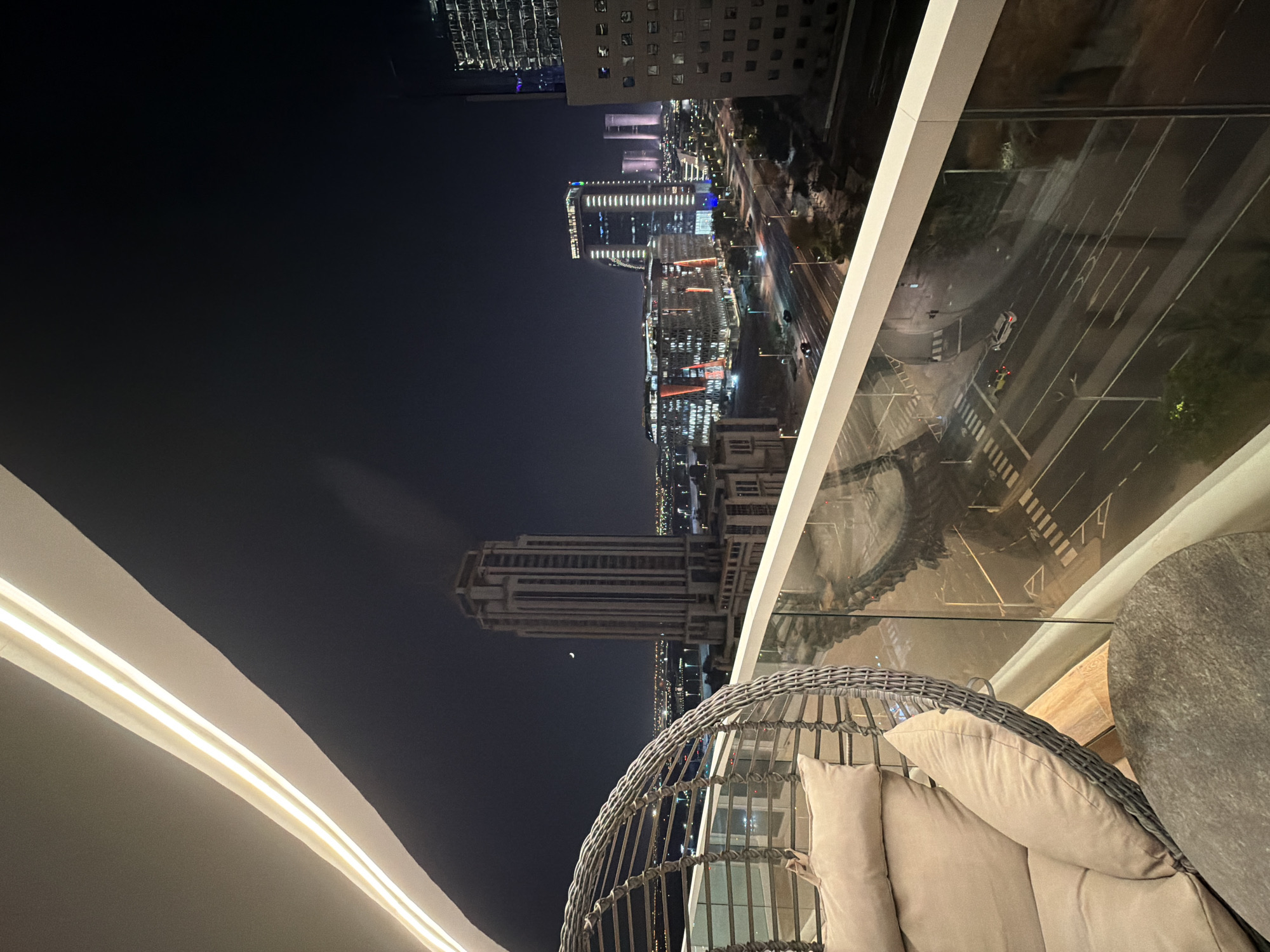 A nighttime aerial view from a balcony shows a city skyline with illuminated buildings and a crescent moon. In the foreground, a woven chair with cushions and a stone table are visible. The balcony railing is made of glass and white metal. Below the balcony, a multi-lane road is visible with cars and streetlights. The city appears to be modern with contemporary architecture.