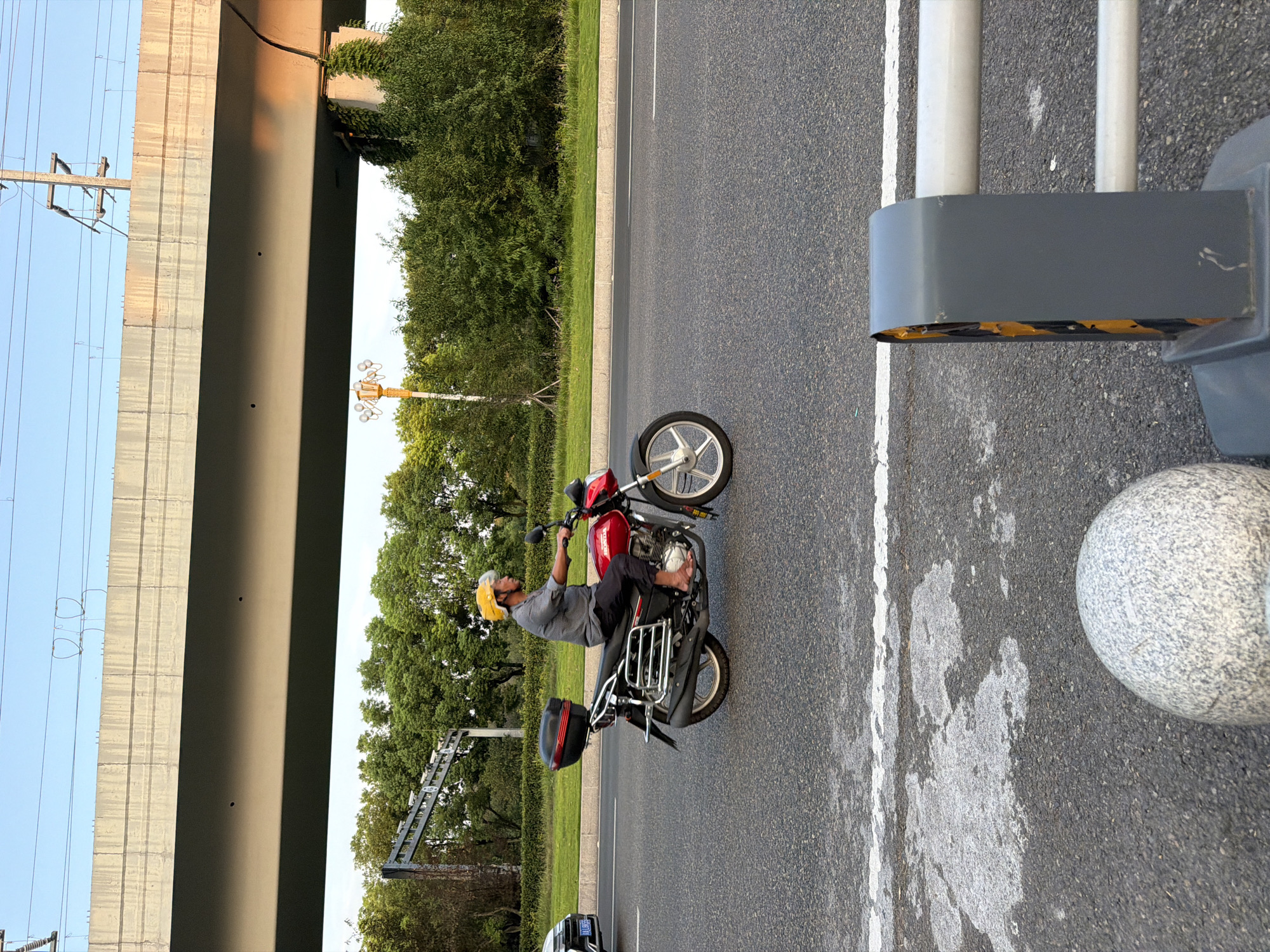 The image depicts a man riding a red motorcycle on a paved road in Jiangjin District, Chongqing, China. The man is wearing a yellow helmet and a grey checkered shirt. The motorcycle has a black storage box on the rear. In the background, there is an elevated concrete structure, possibly an overpass, with electrical wires and poles. Lush green trees and vegetation line the side of the road. The sky is clear and blue, suggesting it is daytime. The foreground shows a portion of the road with white lane markings and a grey traffic barrier.