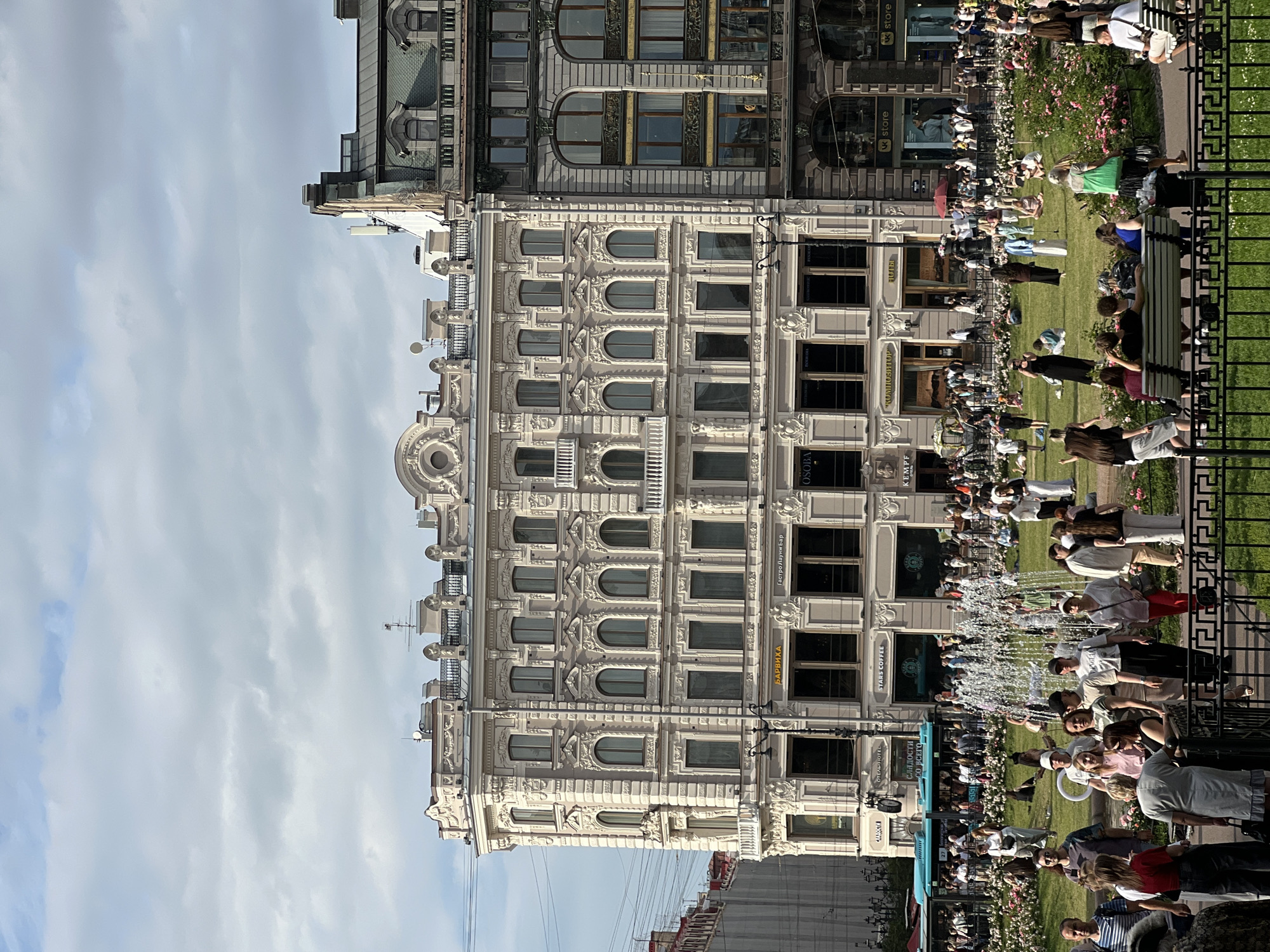This image depicts a sunny day in Saint Petersburg, Russia, likely on Nevsky Prospect. A grand, ornate building with multiple stories and arched windows dominates the center, displaying intricate architectural details. In front of the building, a large park area is filled with people enjoying the weather. Some are seated on benches, others are picnicking on the grass, and many are strolling or standing around. A fountain is active in the middle of the park, with water spraying upwards. A turquoise bus is visible on the left side of the image, suggesting it is a public transportation hub or a busy street. The sky is partly cloudy, but the overall impression is of a pleasant, active day in the city. The buildings appear to house shops and businesses, as indicated by signage like "STARS COFFEE" and "Gastrobar."