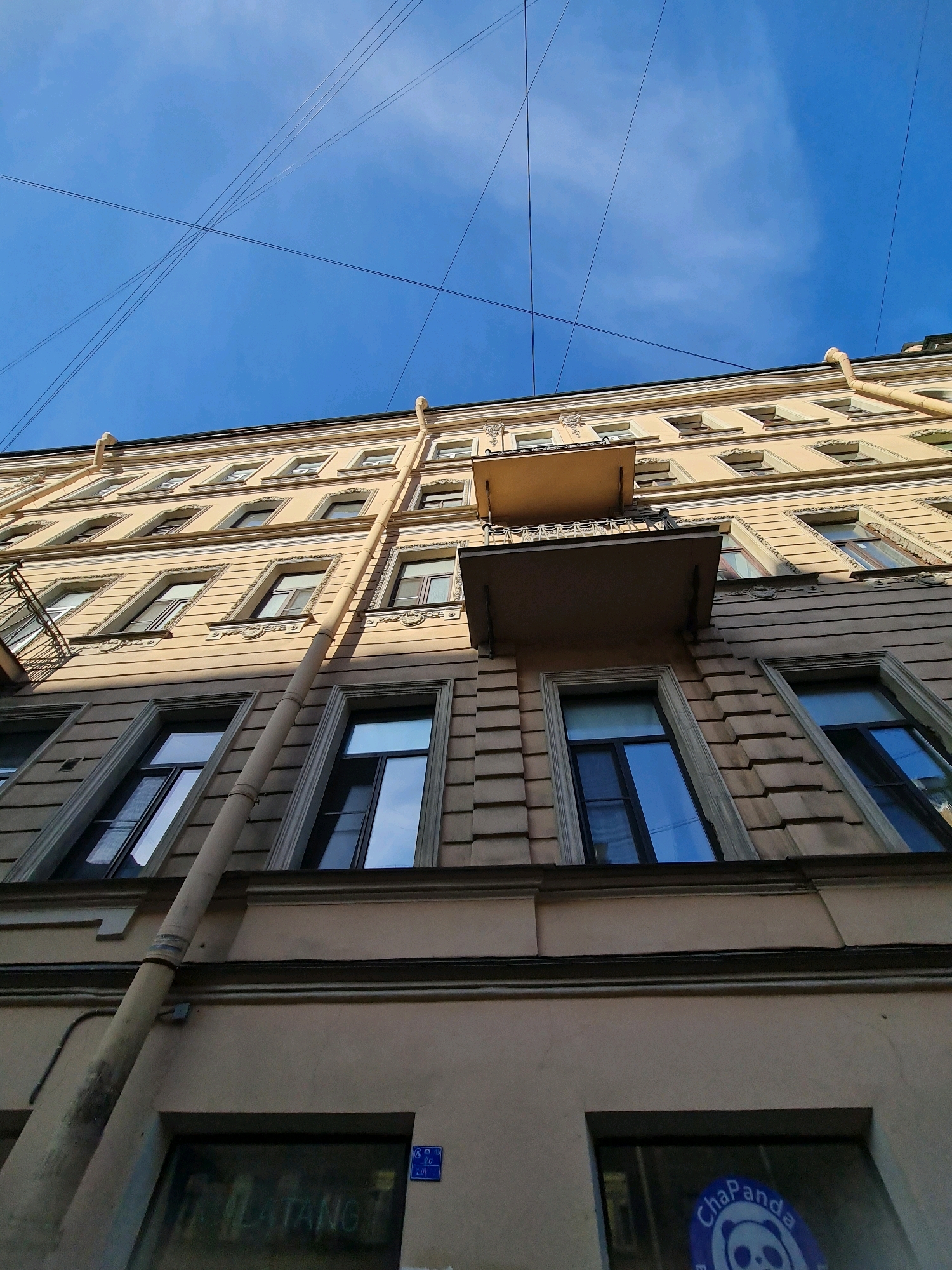 The image shows a low-angle view of a multi-story building facade in Saint Petersburg, Russia. The building is painted a light beige color and features multiple windows with dark frames, some reflecting the blue sky. A prominent cream-colored drainpipe runs vertically down the side of the building. Several thin black wires crisscross the clear blue sky above. On the ground floor, partially visible through large windows, are business signs. One sign reads "ILATANG" in white lettering, and another features a blue circular logo with a panda face and the text "ChaPanda." The architecture suggests a historical building, typical of Saint Petersburg's urban landscape. There are no people visible in the image.