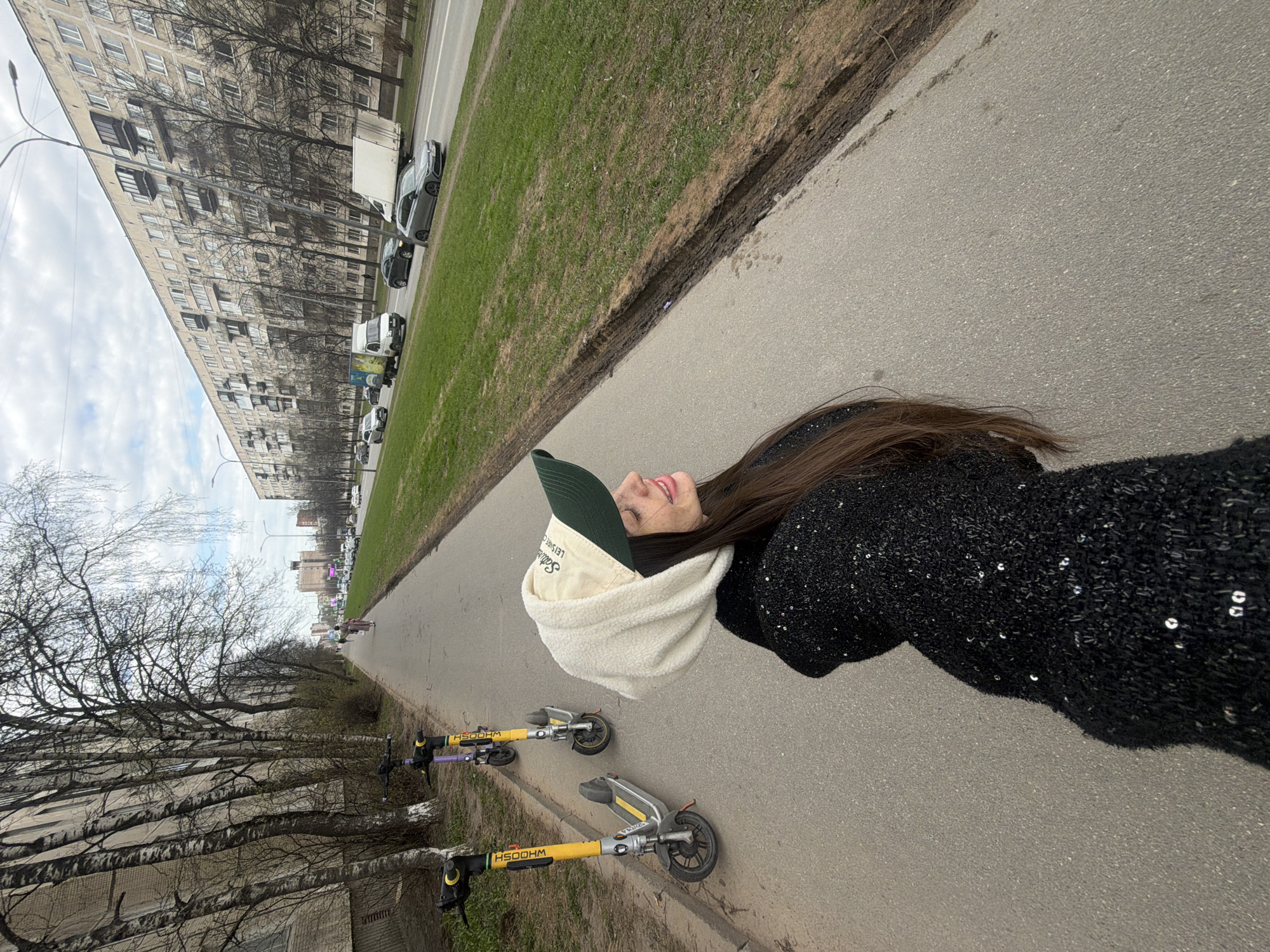 The image displays a sideways view of a person outdoors in an urban setting, likely Saint Petersburg, Russia, given the architectural style. The subject is a person with long brown hair, smiling and looking upwards. They are wearing a dark, possibly sequined, top or jacket, a light-colored fuzzy neck gaiter or scarf, and a dark green baseball cap. The cap has white text that partially reads "Sale Leisure C".
In the background, a very long, multi-story apartment building, characteristic of Soviet-era residential architecture, stretches horizontally. Bare-branched trees, some resembling birches, line a strip of grass between the building and a paved road. Several cars are parked along the roadside.
On the paved surface near the person, two yellow and black electric scooters are parked. The scooters prominently display the branding "HSOOHM" on their vertical handlebars, which, when viewed upright, would typically spell "WHOOSH". The paved area is a sidewalk or pathway, bordered by a strip of grass that shows some muddy or uneven patches, suggesting early spring conditions or recent moisture.
The scene is set during the daytime under an overcast sky, which provides soft, diffuse lighting. The bare trees further indicate either early spring or late autumn. The person's visible smile conveys a positive and cheerful emotion.