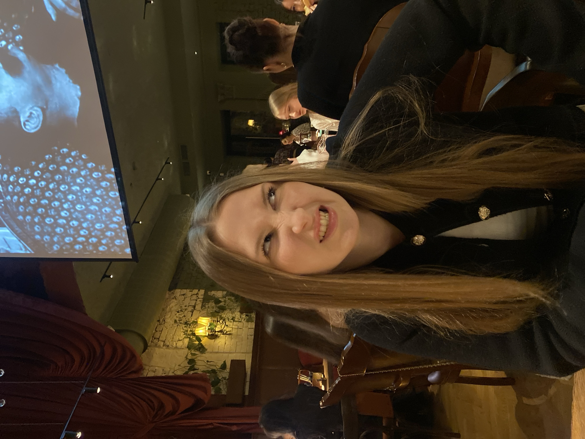 A young woman with long brown hair is posing for a selfie, with an impish grin, in what appears to be a restaurant setting. She is wearing a dark blazer with gold buttons over a white top. In the background, other patrons are seated at tables. There is a projection screen showing a close-up of a face. The restaurant is located in the city of Kazan, Russia, suggested by the interior décor including red velvet drapes, patterned wallpaper, and ornate lighting fixtures.