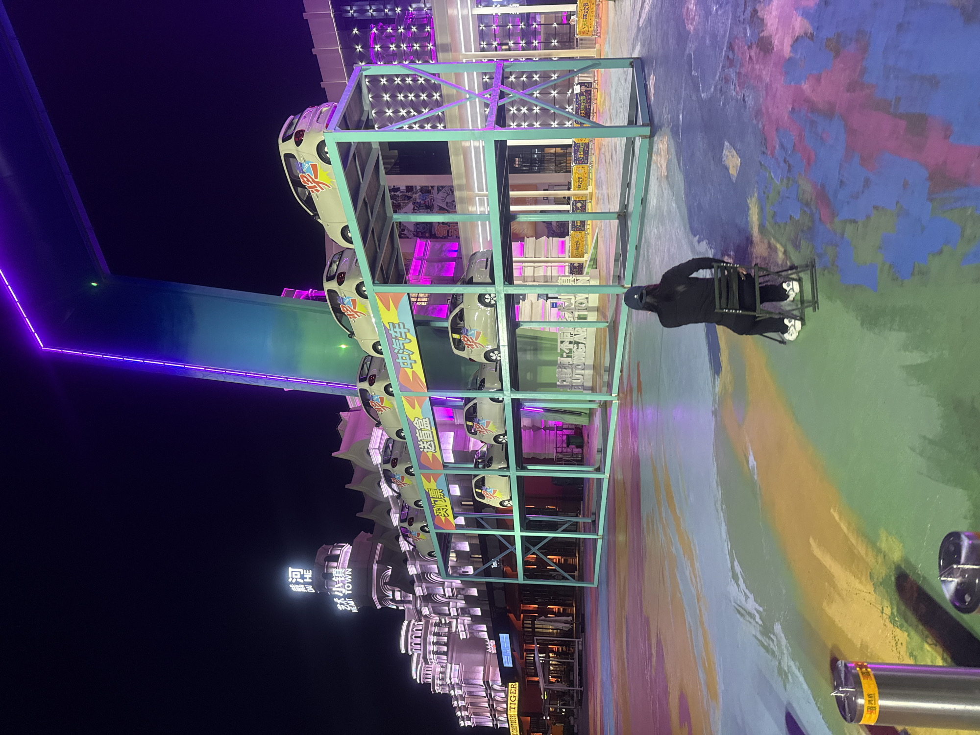 The image depicts a nighttime scene in Qinhuangdao, China, likely at an entertainment or shopping district. In the foreground, a person is seated on a black folding chair facing away from the camera, looking at a colorful, abstract painted ground. The person is wearing a black baseball cap, a black long-sleeved shirt, black pants, and white sneakers.
Behind the person, a multi-level structure displays several miniature white cars, each with colorful decals. This structure is illuminated by pink and purple lights. The background features a modern building facade with intricate architectural details, also lit with pink and purple lighting. Above the building, a large, curved structure with bright purple and blue neon lighting stretches across the sky. Signs in Chinese characters are visible on the building, including "河" and "梦小镇" (Meng Xiaozhen).
The ground is a canvas of vibrant, swirled colors, including shades of green, blue, yellow, pink, and purple, creating an artistic and energetic atmosphere. The overall impression is of a lively urban nightscape with a focus on visual spectacle.