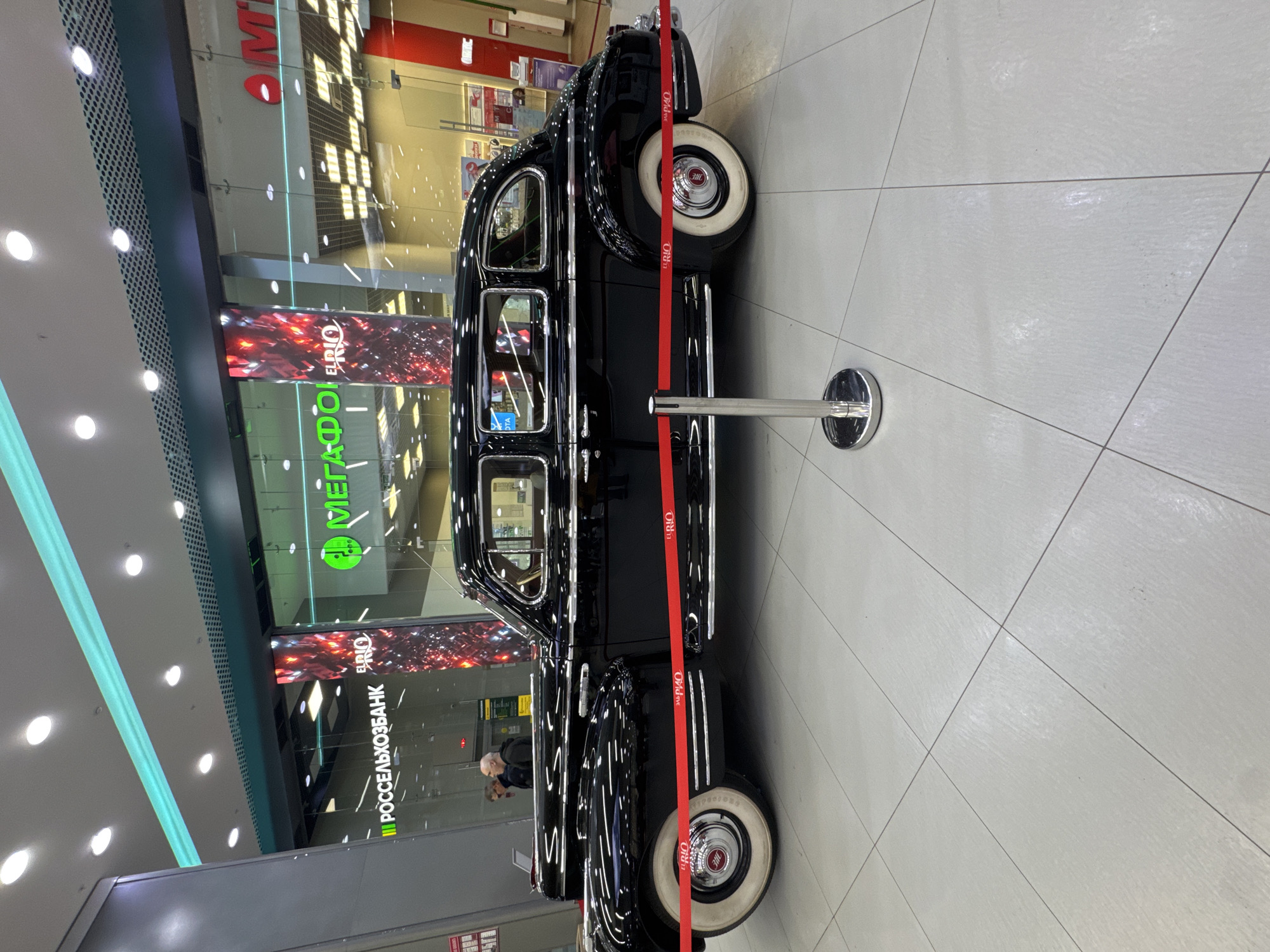 This media file captures a classic black sedan displayed indoors, possibly in a shopping mall or exhibition space, in Samara, Russia. The car, a vintage model, is prominently featured with its polished black exterior reflecting the surrounding lights. A red velvet rope, supported by chrome stanchions, cordons off the vehicle, suggesting it is an exhibit or part of a display.
The background reveals a modern commercial setting. Large glass windows allow glimpses into retail spaces. Visible signage includes Russian text. One sign prominently displays "РОССЕЛЬХОЗБАНК", which translates to "ROSSelkhozbank" (Russian Agricultural Bank). Another illuminated sign shows "МЕТКОФ" and "EL RIO", possibly indicating a store or brand. The overall lighting suggests it is daytime, with artificial lights supplementing natural light filtering in.
A few people are subtly visible in the reflection on the car's windows and in the background, suggesting the presence of shoppers or event attendees. The atmosphere appears calm and observational, with no indication of any specific event beyond the display of the car itself. The pristine condition of the car and the clear demarcation with the rope highlight its status as a point of interest. The tiled floor and modern ceiling lights reinforce the indoor, contemporary commercial environment.