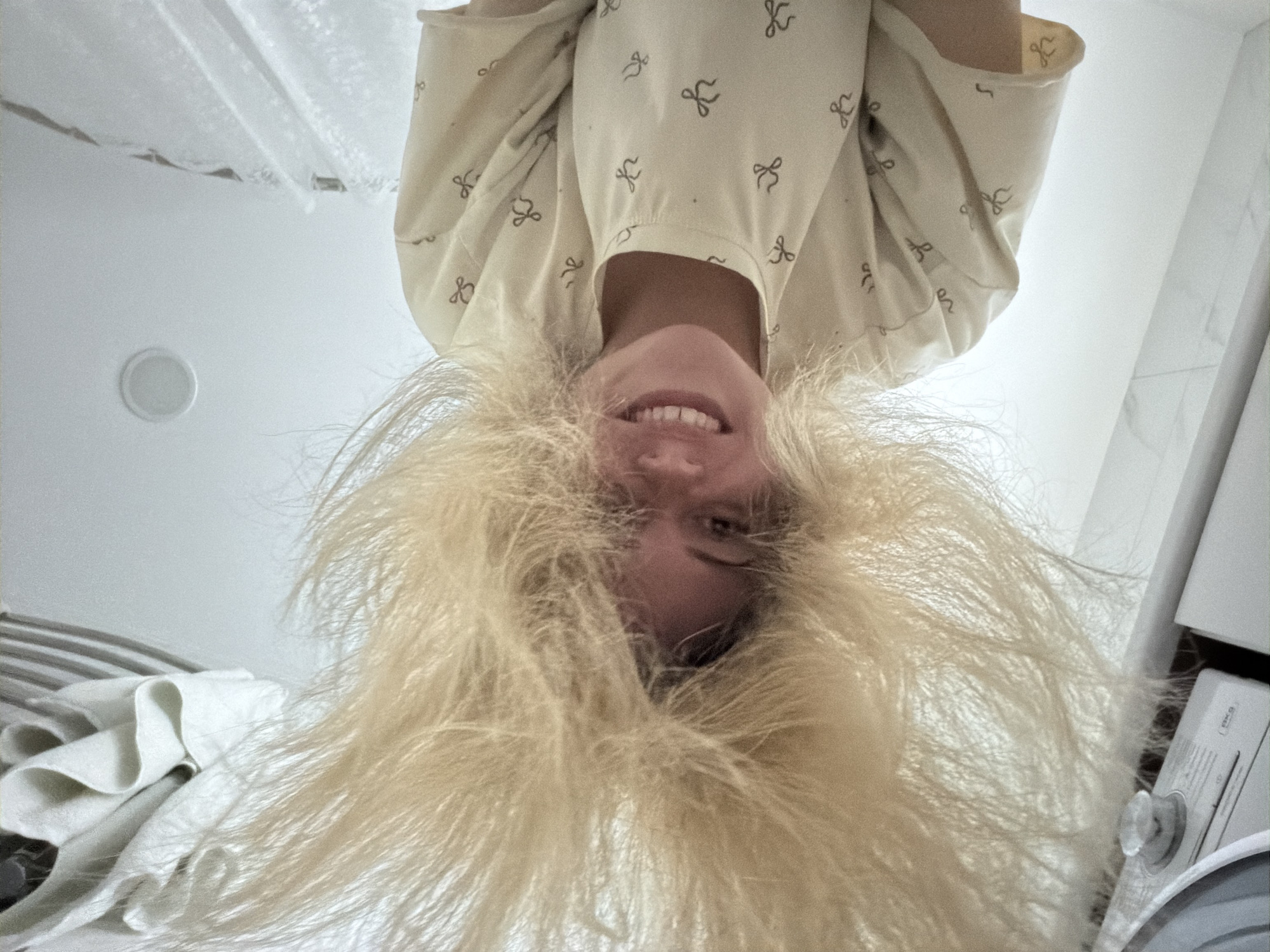This image is a low-angle, upside-down portrait of a person, likely female, looking directly at the camera with a wide, cheerful smile. Their head is inverted, causing their voluminous, bright blonde, fluffy hair to spread out around their face due to gravity. The person is wearing a light-colored top patterned with small, dark, bow-like or knot-like shapes.
The setting appears to be an indoor space, possibly a bathroom or utility area within a residence in Votkinsk, Russia. Various elements of the background support this. To the top left, a white, semi-transparent shower curtain or similar fabric is visible. Below it, on what appears to be a white ceiling from this inverted perspective, a round light fixture is mounted. On the far left, a rumpled white fabric, likely a towel, is draped, with faint stripes visible behind it. On the right side of the frame, a white household appliance, possibly a washing machine or dryer, is partially visible. It has a control knob and some text, with the word "IKEA" distinguishable on a label. The surrounding walls are predominantly white, suggesting a clean, functional space.
The overall mood is playful and joyful, conveyed by the subject's pose, the exaggerated volume of their hair, and their bright, open-mouthed smile. The lighting is soft and even, typical of indoor illumination.