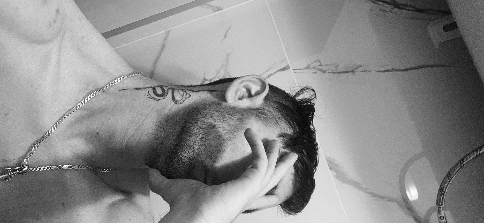 A grayscale photograph shows a person's upper body, head, and hand, with part of a tiled wall in the background. The person is angled with their head tilted to the side. Their left hand is partially covering their face. A silver chain necklace is visible on their chest and neck. There is a tattoo near the hairline above the ear. The background features light-colored tiles with darker veining. The image appears to be taken indoors, likely in a bathroom. The location in the city of Plovdiv, Bulgaria, is not discernible from the image.