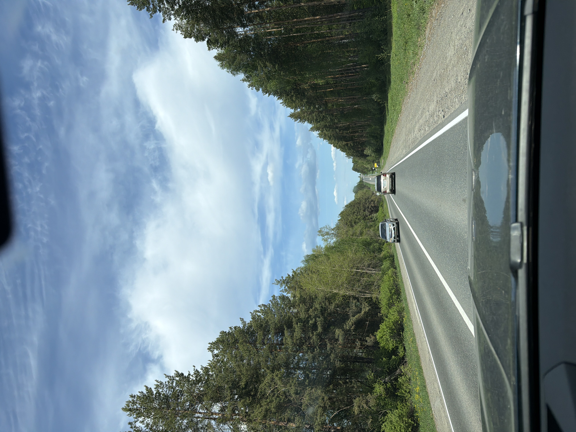 The image shows a view from inside a car, looking out onto a straight asphalt road flanked by tall trees. The sky is visible above the trees and is light blue with scattered clouds. Two cars are visible on the road ahead. The perspective is from the passenger seat of the car. There is no information that allows me to know about the location in the city of Dvigatel', Russia.