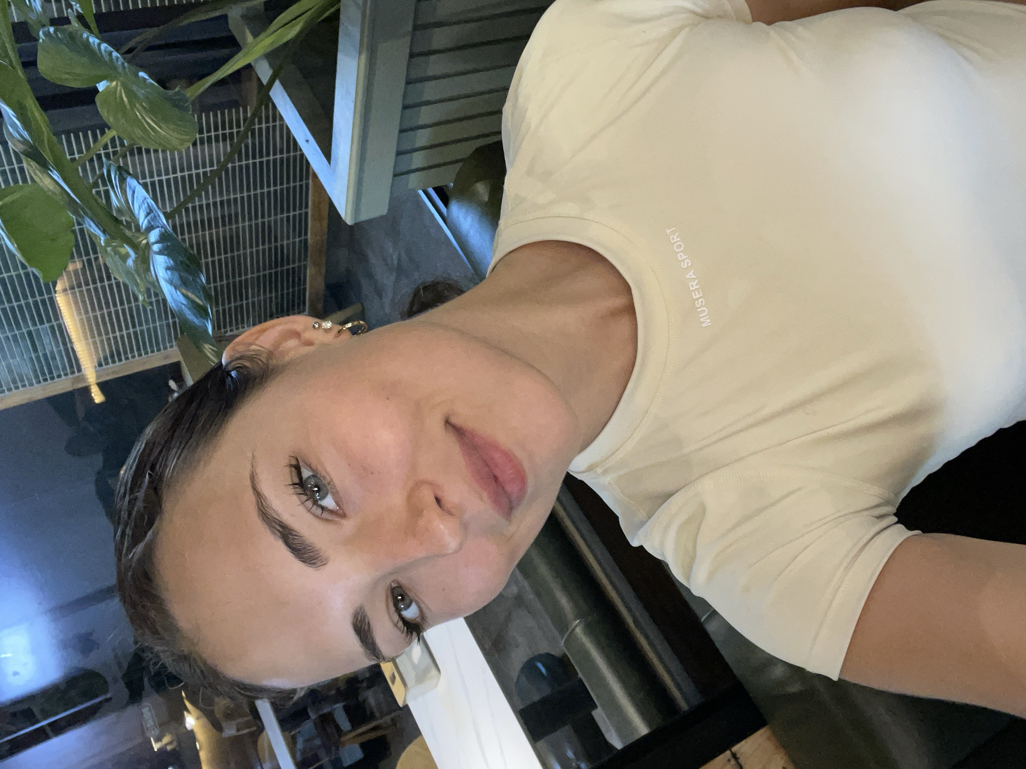 A young woman with brown hair, blue eyes, and light skin is taking a selfie in what appears to be an indoor dining or lounge area. She is wearing a light-colored t-shirt with the text "MUSERA SPORT" printed on the left side of her chest. She has on small earrings and her makeup is subtle, with emphasis on her eyes and lips. The background is blurred, but shows a dark, possibly reflective surface and some greenery from a potted plant. The lighting is dim, suggesting it is evening or the space is dimly lit. The woman is smiling slightly and looking directly at the camera.
