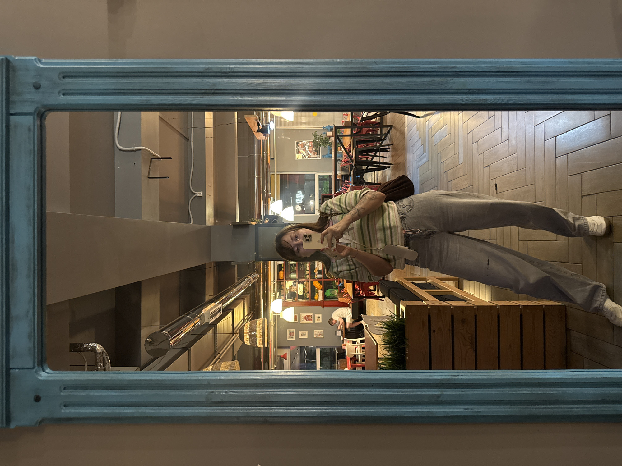 A young woman with brown hair, wearing a striped shirt and loose jeans, stands in front of a large ornate mirror, taking a selfie with her phone. The mirror reflects a cafe interior with wooden floors, tables, chairs, and shelves displaying various items, including pumpkins. In the background, another person sits at a table. The mirror frame is decorative and painted a light blue. The scene appears to be indoors, likely in a cafe or restaurant, and is not specifically identifiable as being in Sochi, Russia.