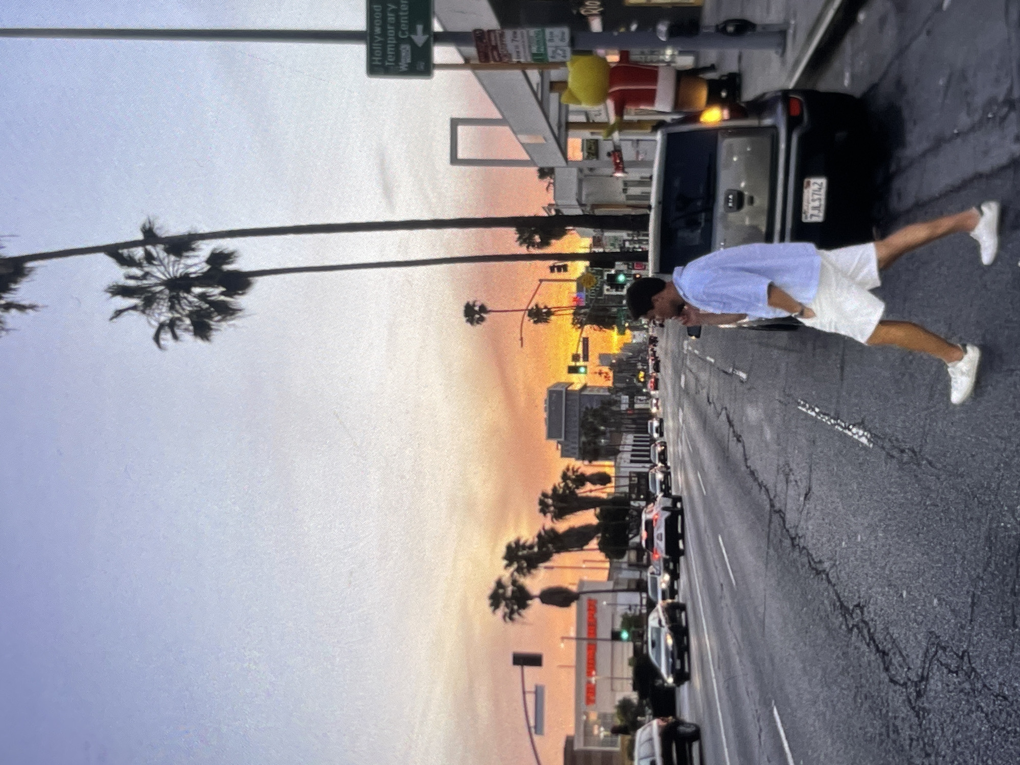 A man is crossing a street in a city at dusk. He is wearing a light blue collared shirt and white shorts. He is walking from right to left in the frame, with his left leg forward. A dark colored car is parked on the right side of the street behind him. In the background, there are palm trees, buildings, and cars on the street. The sky is orange and purple, indicating sunset. A green street sign is visible on the right side of the image.