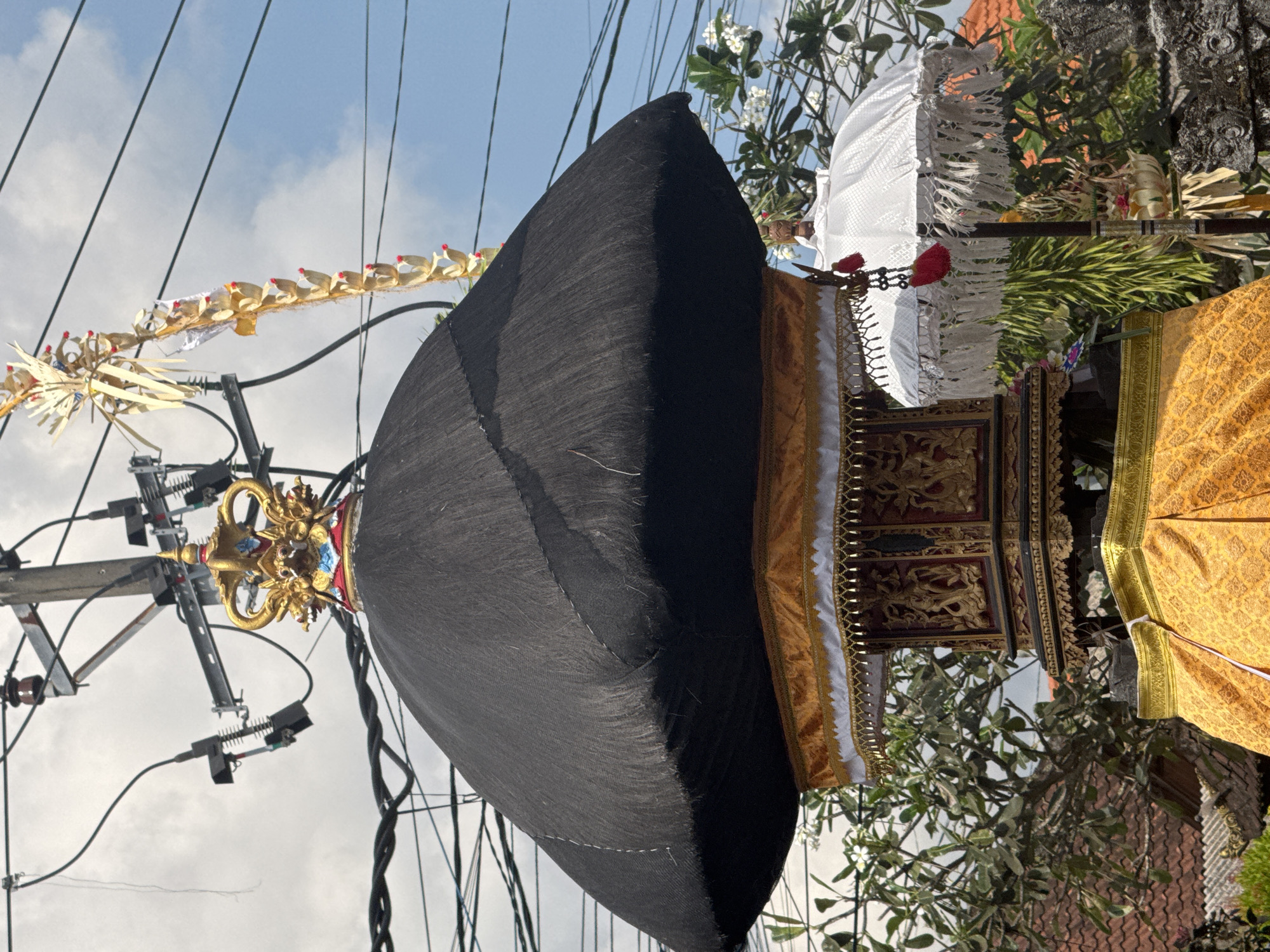 The image shows a religious offering or shrine in the city of Mengwi, Indonesia, set against a backdrop of overhead utility wires and a partly cloudy sky. The primary object is a tiered structure draped in vibrant yellow and gold fabric, topped with a large, dark, mushroom-shaped covering. This covering is adorned with a golden, ornate crown-like finial featuring dragon-like figures. To the right of the main structure, a white, fringed umbrella stands vertically, also decorated with red tassels. Green foliage and a section of a tiled roof are visible in the background, suggesting an outdoor setting. The overall scene suggests a traditional cultural or religious event.
