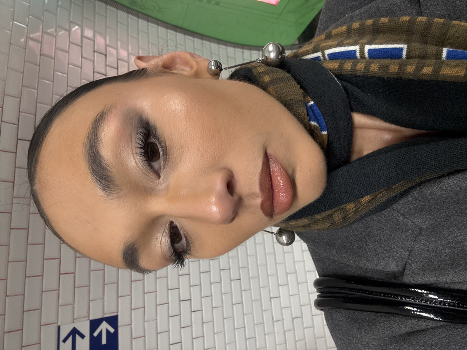 A close-up, chest-up image depicts an individual with a medium-dark complexion, prominent eyebrows, and full, glossy lips. The subject wears extensive dark eye makeup. Attire includes a gray coat or blazer and a multi-patterned scarf featuring dark blue, brown, and white geometric designs. Silver spherical drop earrings are visible on both ears, with a shiny black strap (likely a bag strap) over the left shoulder. The background reveals a wall constructed of white rectangular tiles with red grout lines, characteristic of the Paris Métro system. Two blue rectangular signs with white left-pointing arrows are affixed to this tiled wall, indicating direction. To the right, a curved green structure and a subtle pink light source are present. The individual is positioned slightly angled, looking downwards and to the right of the camera.