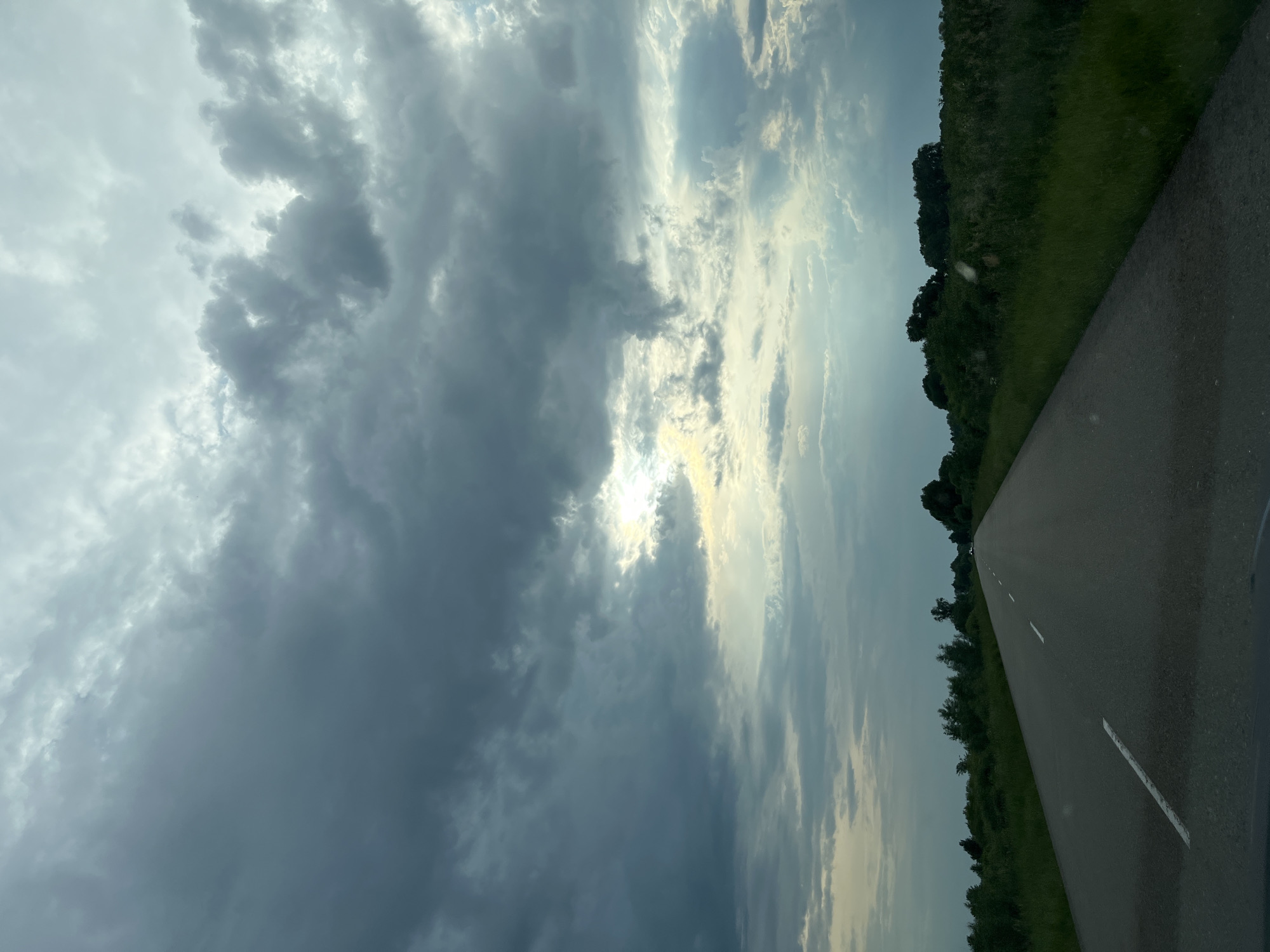The image depicts a straight asphalt road extending into the distance through a rural or semi-rural landscape in Pohreby, Ukraine. The perspective is from within a vehicle, looking forward. The sky is largely overcast, dominated by dark grey and lighter grey cloud formations. A bright area on the upper right-mid portion of the sky indicates the sun partially breaking through the clouds, casting diffuse light and some visible light rays.
The two-lane road features white dashed lane markers. The immediate foreground shows the road surface and a slight dark reflection consistent with a vehicle's dashboard. On both sides of the road, dense green vegetation, including tall grass, bushes, and trees, lines the verges and fields. The vegetation on the right appears thicker and taller than on the left. In the far distance, a dark line of trees defines the horizon. A light-colored vehicle is faintly visible on the road in the far distance, receding from the viewer. No individuals are present in the visible portion of the scene. The lighting suggests an overcast condition, possibly during late afternoon or early morning, with the sun attempting to emerge from behind the clouds.