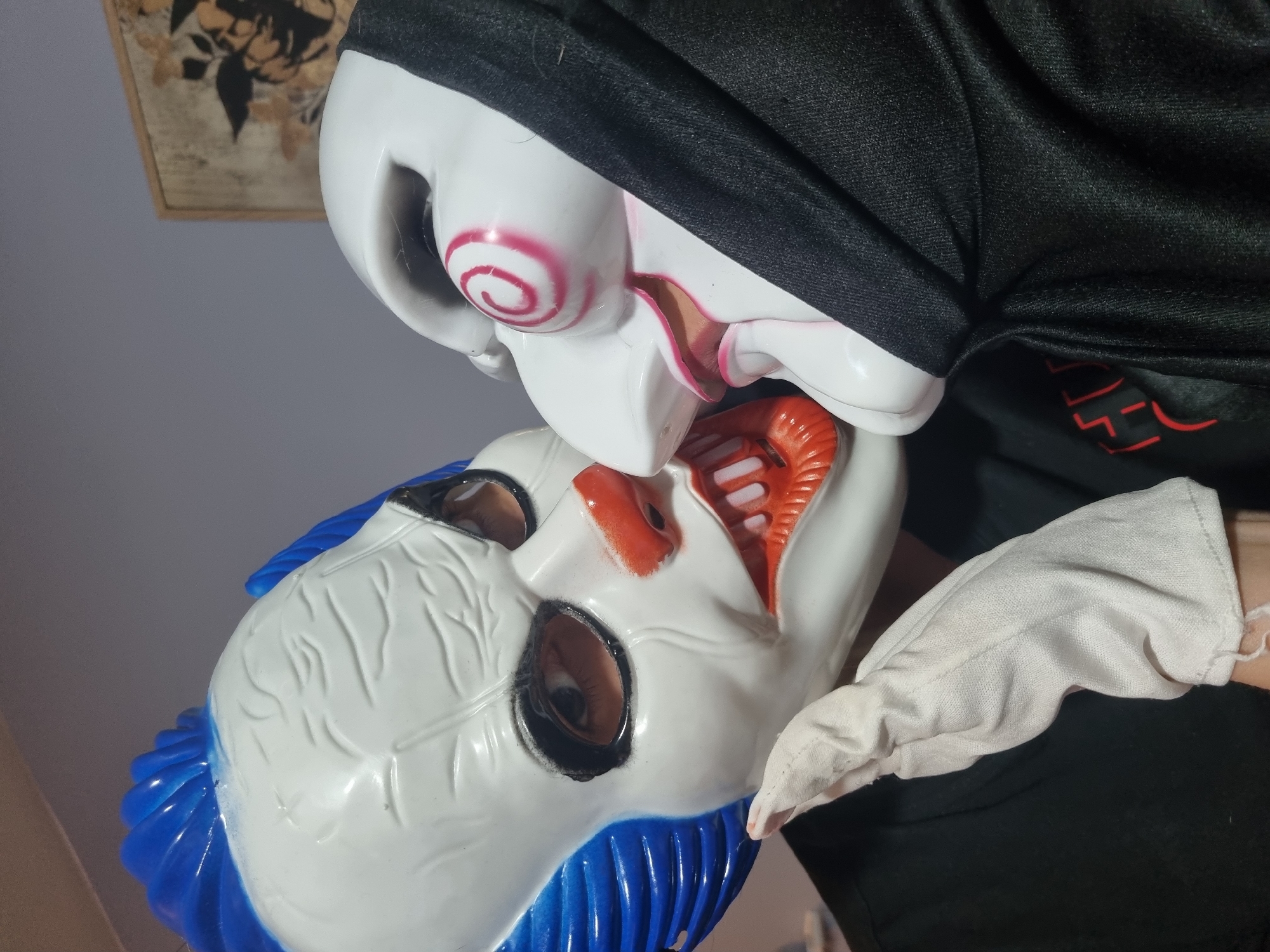 A close-up shot features two Halloween masks in an indoor setting. The upper mask, primarily white with a black hood, has a red spiral design on its cheek and exaggerated facial features. The lower mask, also white, has blue hair and an orange mouth with pronounced teeth. A person wearing a black shirt and a white glove appears to be holding the masks. The background is blurred, but a framed artwork is partially visible in the upper left corner. The location is undetermined.