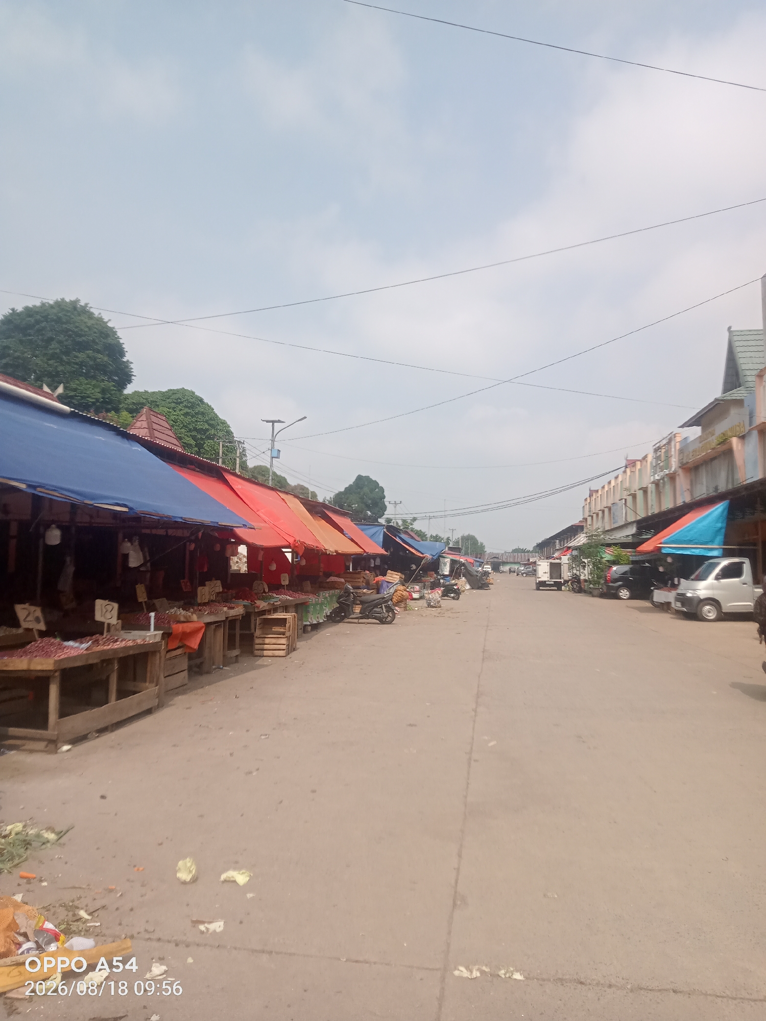 This is a photograph of an outdoor market scene in Jambi, Indonesia, taken on August 18, 2026, at 09:56. The market appears to be a street market with stalls set up along a paved road. On the left side of the road, there are numerous stalls covered by blue and red tarps, displaying goods on wooden tables. Some of these tables hold what appear to be produce, such as onions and other vegetables, with some items in wooden crates. There are also price tags visible on some of the tables. A few motorcycles are parked in front of the stalls. Further down the road, more stalls and buildings are visible, with a variety of vehicles, including cars and a white truck, parked. The right side of the road features a row of more permanent buildings with awnings, also housing market stalls. The sky is partly cloudy. The overall scene depicts a bustling commercial area typical of a local market in Indonesia.