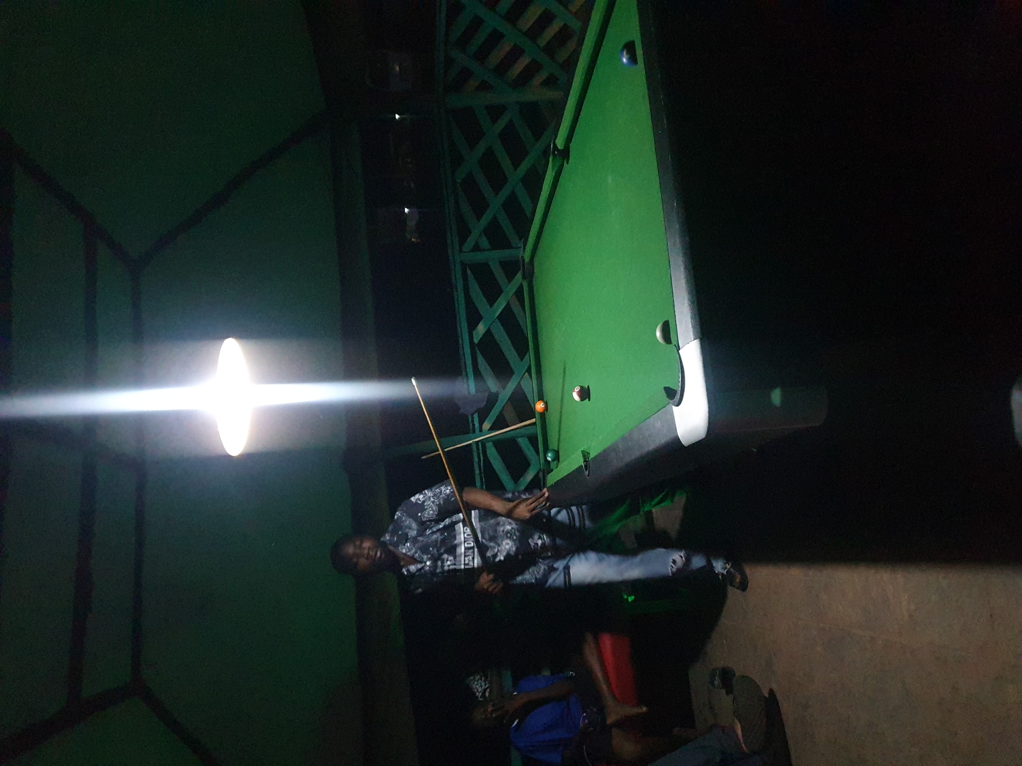 The image depicts an indoor setting, likely a recreational area, with a pool table as the central object. The lighting suggests it is nighttime. Three individuals are visible, though their faces are not clearly discernible in all cases due to the low light conditions. One man, wearing a patterned shirt and jeans, stands near the pool table, holding a cue stick, appearing to be playing or about to play. Another person is seated on a red chair, wearing a blue shirt. A third individual is partially visible in the background, also seemingly seated. The walls of the room are painted green, and there is a structure with a latticed design in the background, possibly a roof or partition. The overall atmosphere is casual and relaxed, typical of a gathering for a game of pool. There is a bright light source hanging from the ceiling, casting a strong glare. No visible text is present in the image.