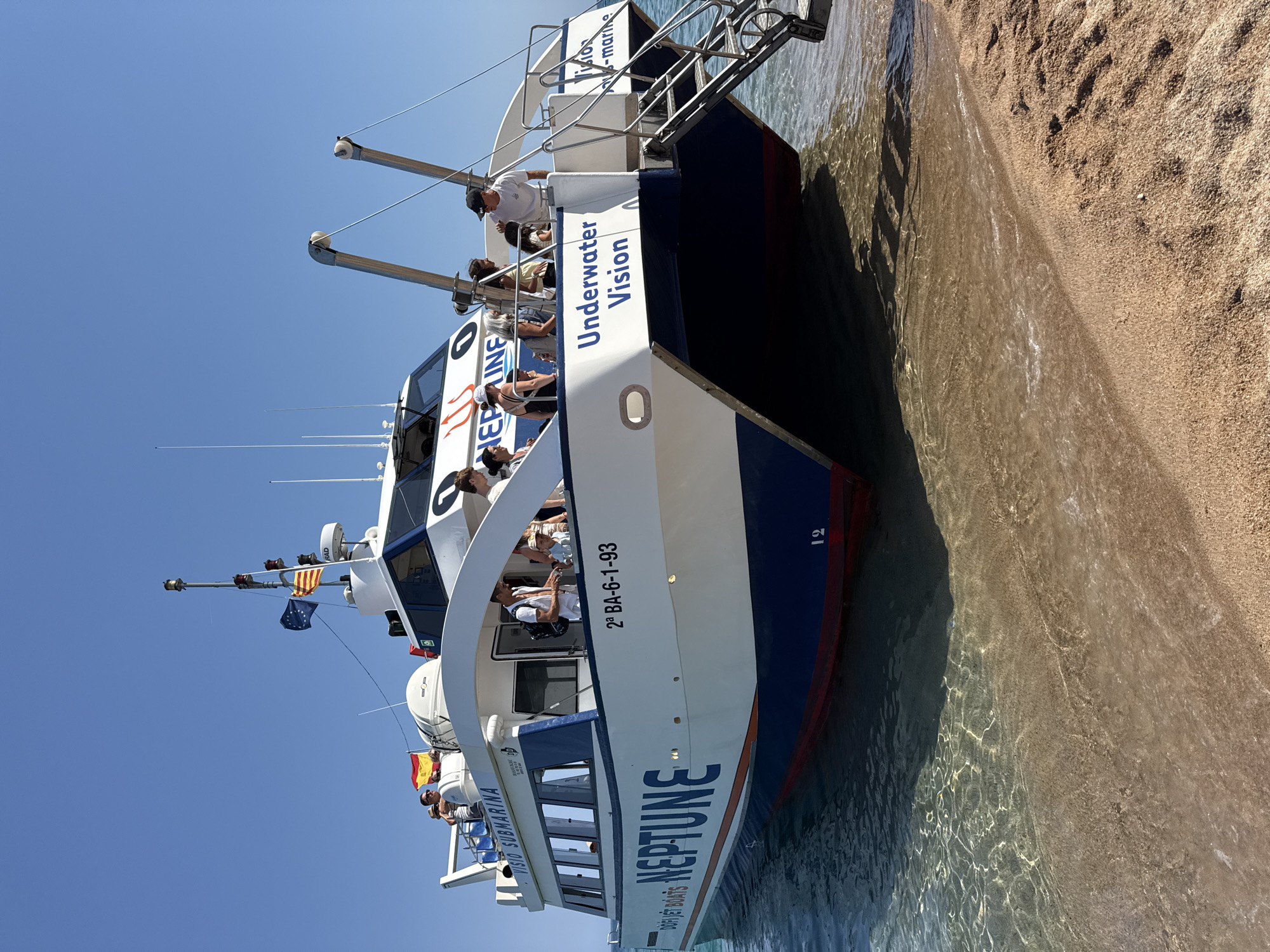 This is a photograph of a boat named "NEPTUNE" docked at a sandy shore. The boat appears to be a tourist vessel, possibly a glass-bottom boat or submarine tour given the text "VISION SUBMARINA" visible on its side. Several people are on the deck of the boat, and some are looking towards the shore or the water. The boat is white with blue trim, and the name "NEPTUNE" is prominently displayed in large blue letters. A Spanish flag and other maritime flags are also visible on the boat. The water is clear and shallow, lapping at the sandy beach, and the sunlight creates dappled patterns on the water's surface. The sky is clear and blue. The overall scene suggests a sunny day, likely in the late morning or early afternoon, and the atmosphere appears relaxed and leisurely. Text visible on the boat includes "NEPTUNE," "VISION SUBMARINA," "UNDERWATER VISION," and "2ª BA-6-1-93." The location is identified as Blanes, Spain.