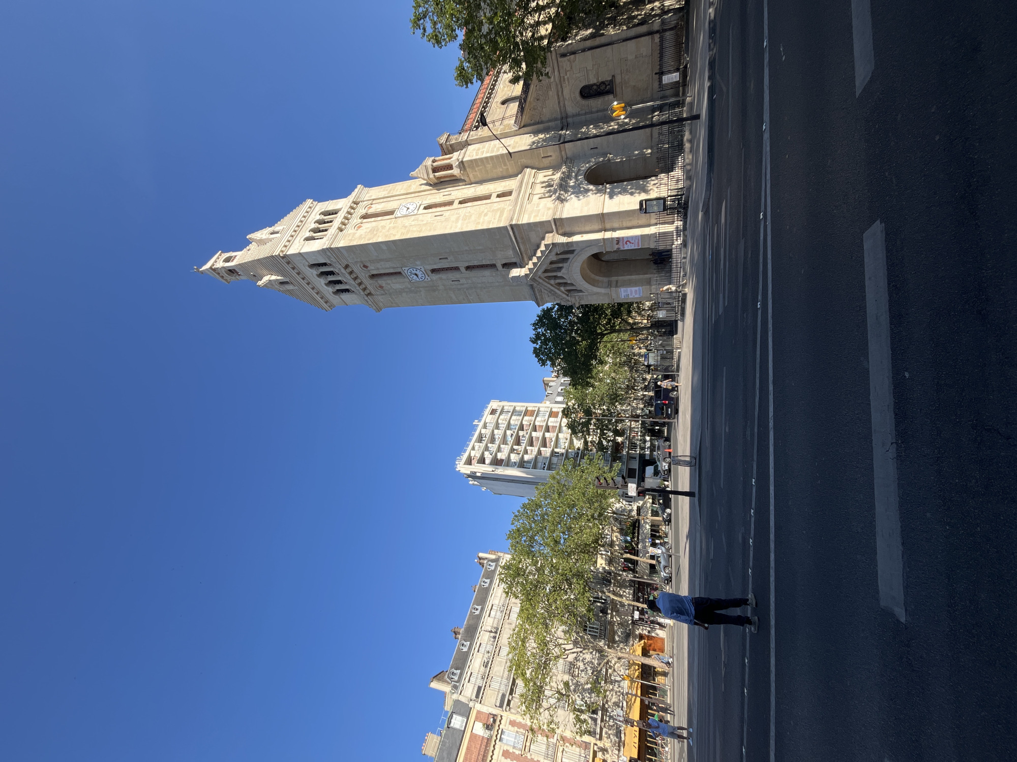 The image depicts a street scene in Paris, France, featuring the Church of Saint-Charles de la Butte-Chaumont. A tall, light-colored bell tower of the church dominates the right side of the frame, its architectural details illuminated by sunlight against a clear blue sky. In the foreground, a wide street with crosswalk markings is visible, with a man in a light blue shirt and dark pants standing near the center, facing away from the camera.
To the left of the church, a row of Parisian apartment buildings with balconies and ornate facades lines the street. Trees with green foliage are scattered along the sidewalk and in front of the buildings, providing shade. Several cars are parked and in motion on the street, and pedestrians are visible walking on the sidewalks. A yellow awning of a business can be seen on the left side of the image. A yellow and red Metro sign is mounted on a pole near the church entrance. The overall impression is a sunny day in an urban Parisian setting.