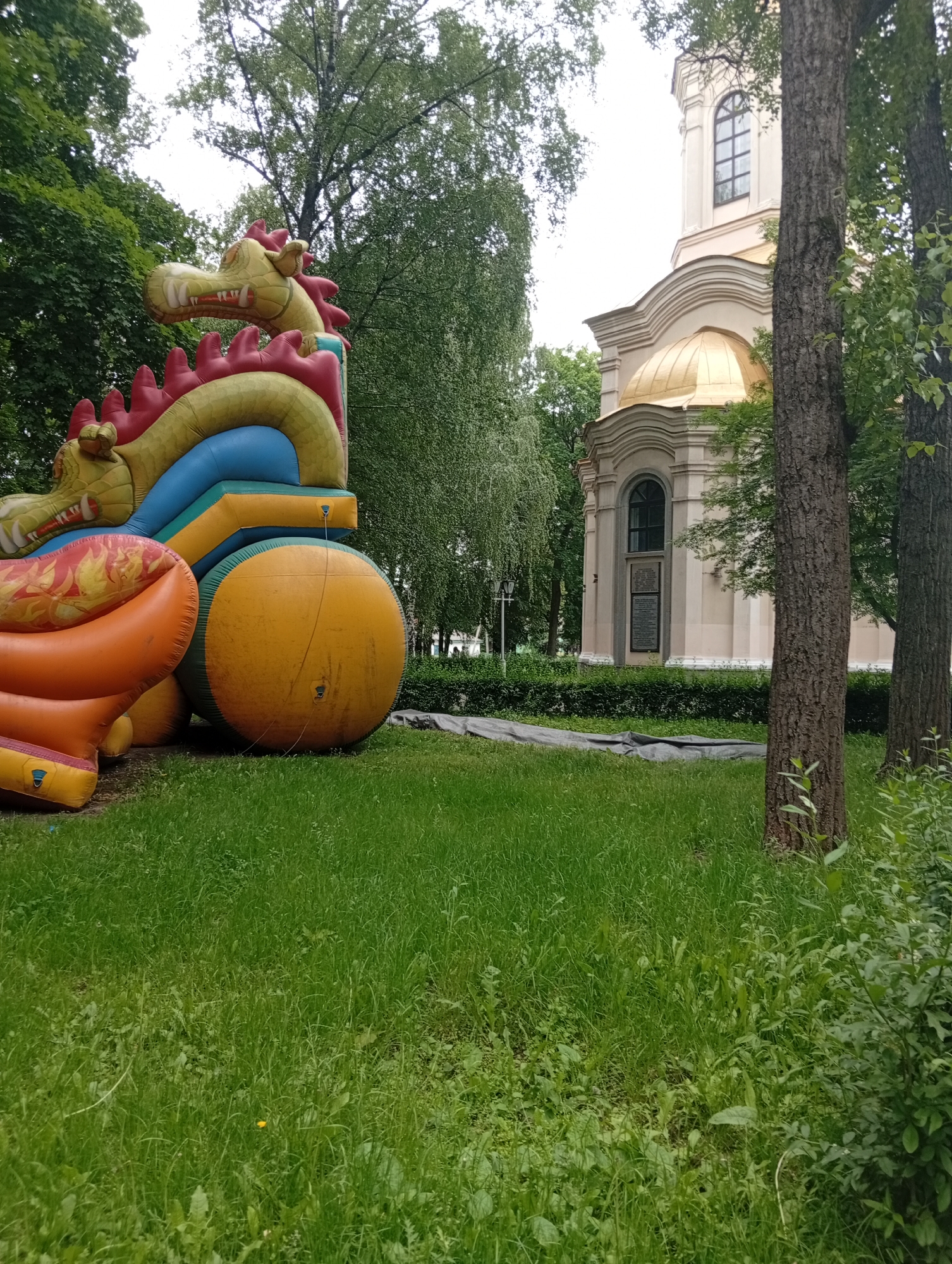 The image shows an inflatable dragon toy in the foreground, with a church in the background. The dragon is yellow and red, with blue and green accents. It appears to be partially deflated. The church is white with a gold dome. The scene is set in a park with lush green grass and trees. The overall impression is one of a sunny day in an urban park, with children's entertainment set up near a historical building. The specific location in Poltava, Ukraine, is not discernible from the image itself, but it is likely a public park or recreational area.