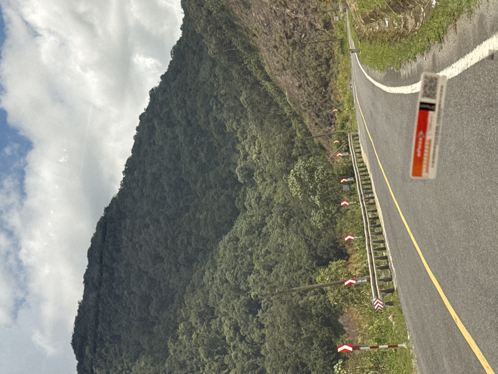 The image depicts a winding asphalt road cutting through a mountainous landscape covered in dense green forest. The road is bordered by a metal guardrail and marked with a yellow lane divider. Several red and white chevron signs are visible along the roadside, indicating a curve in the road. The sky above is partially cloudy, with patches of blue visible between the white clouds. In the foreground, a blurry sticker with red and white markings and a QR code is visible, affixed to the road surface. The overall scene suggests a scenic route in a rural or mountainous area, likely a highway or provincial road in Vietnam, given the context of Nam Khánh Vĩnh.