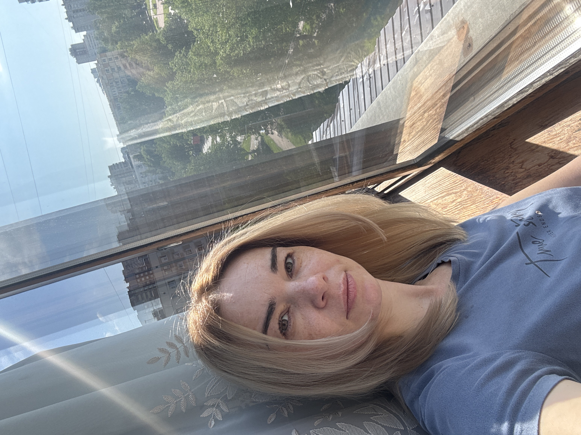 A blonde woman is lying on a patterned, blue-grey surface, potentially a couch or a bed, wearing a light blue t-shirt with dark writing. The photo seems to be taken from a slightly above angle. Behind her, there is a large window with a reflection of city buildings and trees, suggesting a daytime setting. The location is Unknown, Russia. The window frame is visible with a wooden interior and screen.