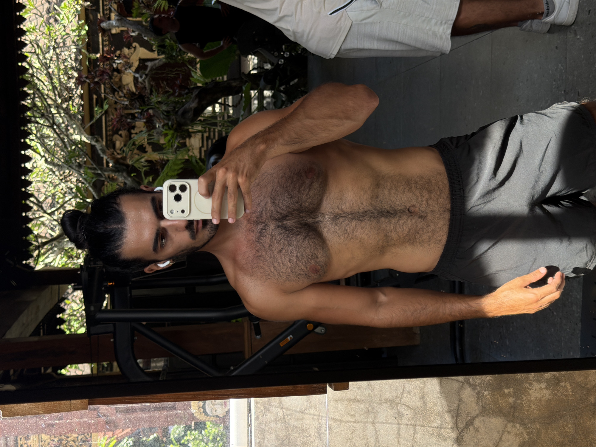 A man is reflected in a mirror, shirtless and wearing black shorts, taking a selfie with a white iPhone. He has dark hair pulled back in a bun and is wearing an earpiece. The reflection also shows a person in the background wearing white shorts and a black top. Outside the reflection, there is greenery and a wooden structure, suggestive of an outdoor setting in North Kuta, Indonesia.